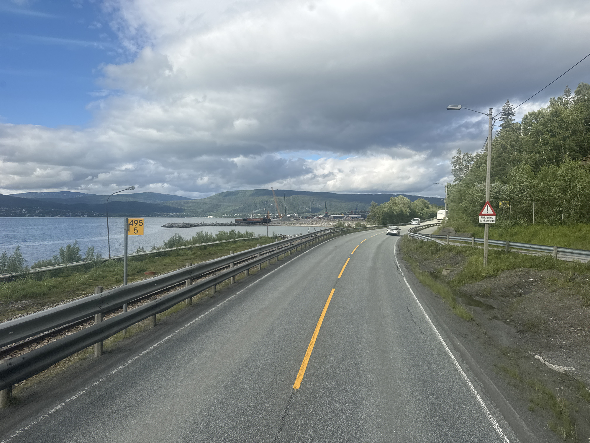 The image depicts a scenic road in Mo i Rana, Norway, with a body of water to the left and a forested hillside to the right. The road is asphalt with a yellow center line and a white edge line. A guardrail runs along the left side of the road, separating it from the water. In the distance, mountains are visible across the water, and there is some industrial activity with cranes. A yellow sign with the numbers "495 5" is posted near the roadside. Further down the road, a white car is visible. On the right side of the road, a triangular warning sign with a red border and a black exclamation mark is posted, with text below that reads "Utkjøring tankanlegg." The sky is filled with dramatic clouds, suggesting a dynamic weather system. The overall impression is one of a coastal highway winding through a natural landscape with signs of human industry in the distance.