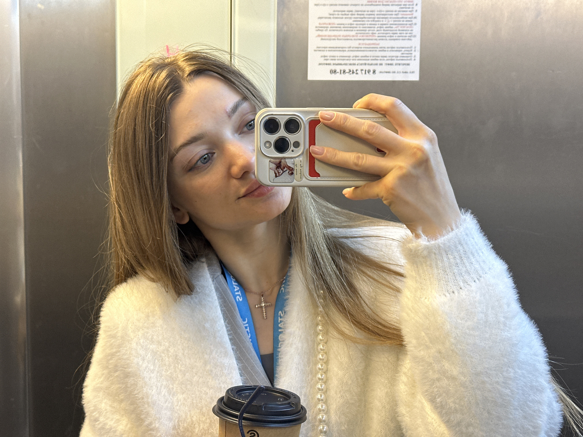 A young woman is taking a selfie in what appears to be an elevator. She is holding an iPhone in her right hand, and the phone's case has a picture of a cat on it. The woman has blonde hair, blue eyes, and light skin. She is wearing a white, fuzzy sweater, a blue lanyard, and a necklace with a cross pendant. She also has a string of pearls draped around her neck. In her left hand, she holds a disposable coffee cup with a black lid. The background shows the interior of the elevator, with metal walls and a sign with text in Russian, possibly indicating the location is Kazan, Russia. The lighting is bright and slightly diffused.