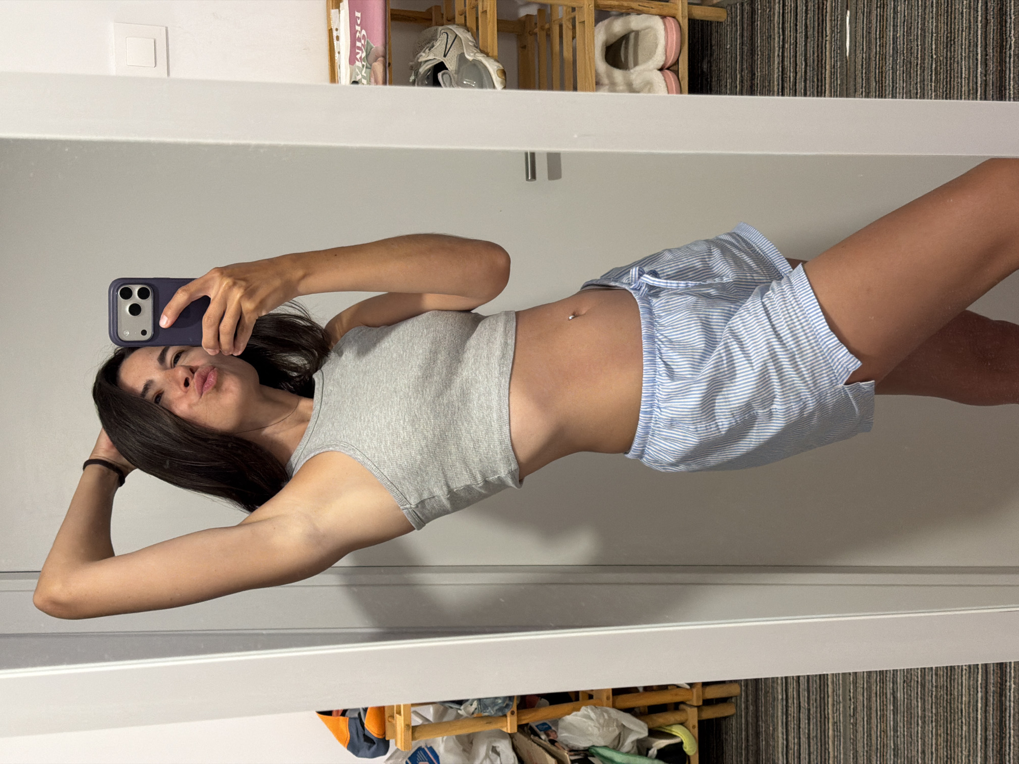 A young woman with dark hair is taking a mirror selfie in a brightly lit room. She is wearing a grey ribbed crop top and blue and white striped pajama shorts. Her left arm is raised behind her head, and she is puckering her lips. Her belly button is pierced. In the background, to the left, is a wooden shelving unit with various items, including shoes and bags. To the right, a white door with a silver handle is visible, and a white light switch is mounted on the wall. A small shelf unit with shoes and a pink book is also visible on the right. The flooring appears to be carpet with a striped pattern. The scene is indoors, likely a bedroom or dressing area, located in L'Hospitalet de Llobregat, Spain.