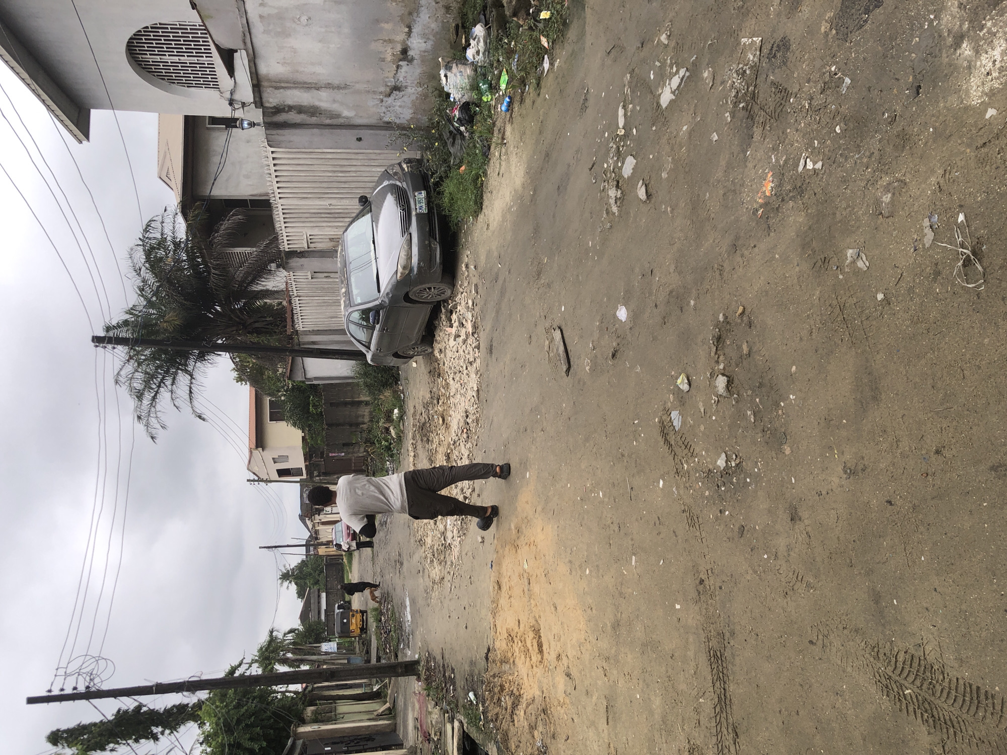 In Ado, Nigeria, a person wearing a white t-shirt and dark pants walks away from the camera on a dirt road. The road is littered with debris, including rocks, sand, and various types of trash. In the background, a gray sedan is parked on the side of the road next to a white fence. A yellow tricycle is also visible further down the road, along with other people and buildings. The sky is overcast, and palm trees are scattered throughout the scene. The overall impression is one of an urban environment with significant issues of waste management.