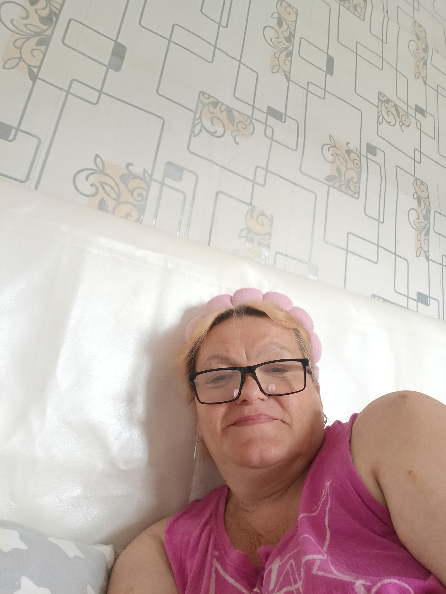 A woman with short, light hair is lying on a white headboard or padded surface, possibly a bed or couch. She is wearing black-rimmed glasses and a pink hair accessory that resembles small pom-poms or flowers. She is wearing a pink tank top with a white design. She has light skin and is looking at the camera with a slight smile. The background features wallpaper with a geometric design and floral patterns in beige and grey. The setting appears to be indoors, likely a bedroom. There is no specific indication that the image was taken in the city of Красный Маяк, Russia.