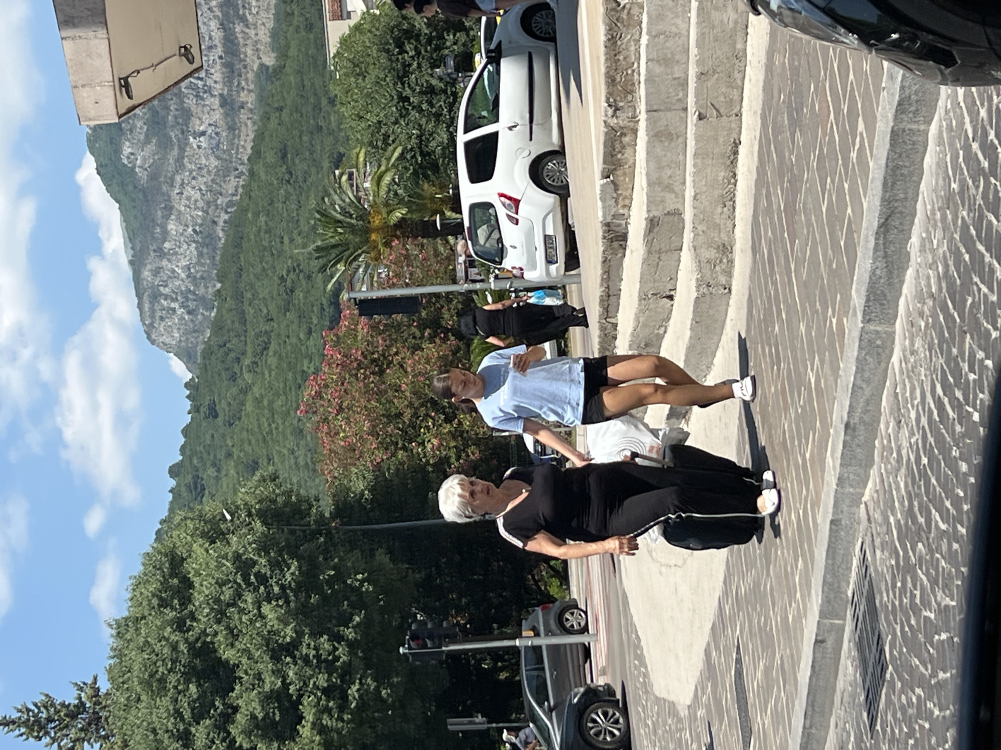 The image shows a street scene in Budva, Montenegro, on a sunny day. Two women are the main subjects, standing on a paved area with stairs to their right. The woman in the foreground, with white hair, wears a black outfit with white stripes on the sleeves and down the sides of her pants. She is looking towards her left, with a slight smile. The second woman, behind and to the left of the first, wears a light blue t-shirt and dark shorts. She is looking down and to her right.
Behind the women, a white car is parked. Further in the background, lush green trees and a rocky mountain with some vegetation are visible under a blue sky with scattered clouds. The paving stones are a notable detail, laid in a diamond pattern. The scene suggests a casual, daytime activity, possibly a walk or break during a visit. There are no other discernible activities or events taking place beyond the presence of the individuals and the parked vehicle. The lighting indicates it is daytime, and the clear sky suggests fair weather. The overall impression is a bright and pleasant outdoor environment. No visible text is present in the image.