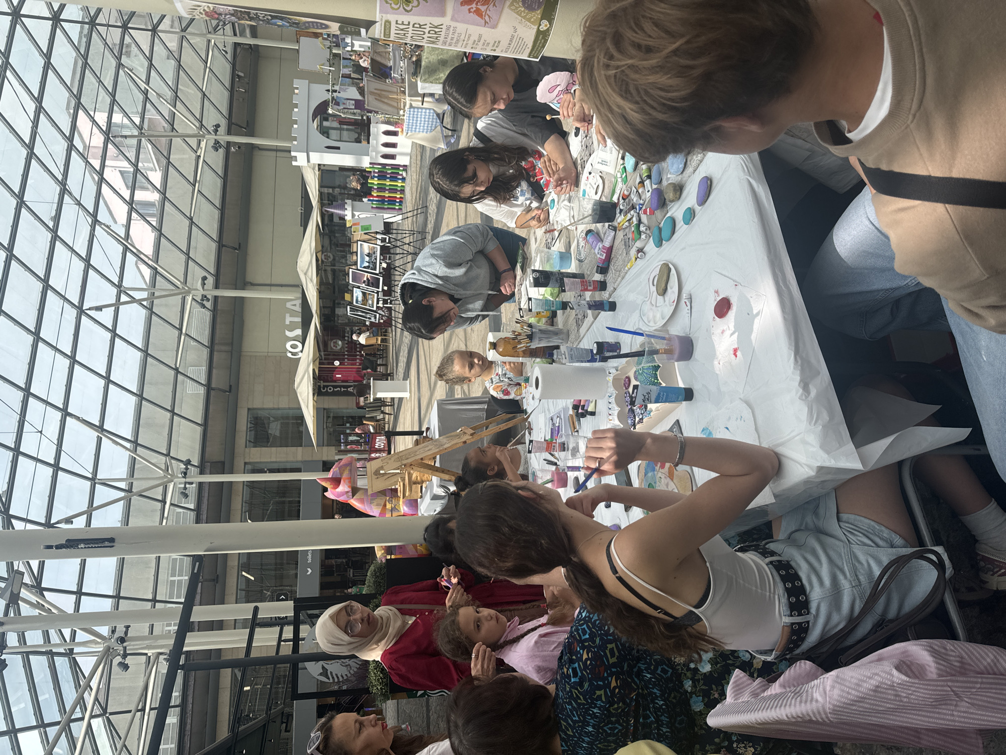 This image depicts an outdoor art workshop taking place in a covered walkway or shopping area with a glass roof. The setting suggests it could be a public space within the city of Kilkenny, Ireland, possibly an area like the Kilkenny Medieval Mile, which features shops and public spaces.
In the foreground, a group of individuals, predominantly women and girls, are gathered around a long table covered with a white paper tablecloth. They are engaged in painting, with various art supplies scattered across the table, including paint tubes, brushes, water cups, and palettes. Some people are actively painting on rocks, while others appear to be observing or preparing their materials.
Several people are looking down at the table, focused on their artistic endeavors. One woman, wearing a red top and a hijab, stands behind the table, holding paintbrushes. To her left, a woman in a black and blue patterned top is seated. A young girl with braided hair in a pink top is seated at the table, looking towards the center.
In the mid-ground, more people are visible around the table, including several young women and girls painting. Behind them, the environment opens up to reveal more stalls or shops with umbrellas and awnings. There are framed pictures displayed on easels, suggesting other art-related activities or a market. A small, white castle-like structure is also visible in the background, possibly a decorative element or part of a children's play area. The overall atmosphere seems to be one of a community event or workshop in a busy, open-air commercial or pedestrian area. The lighting suggests it is daytime, with sunlight filtering through the glass roof.