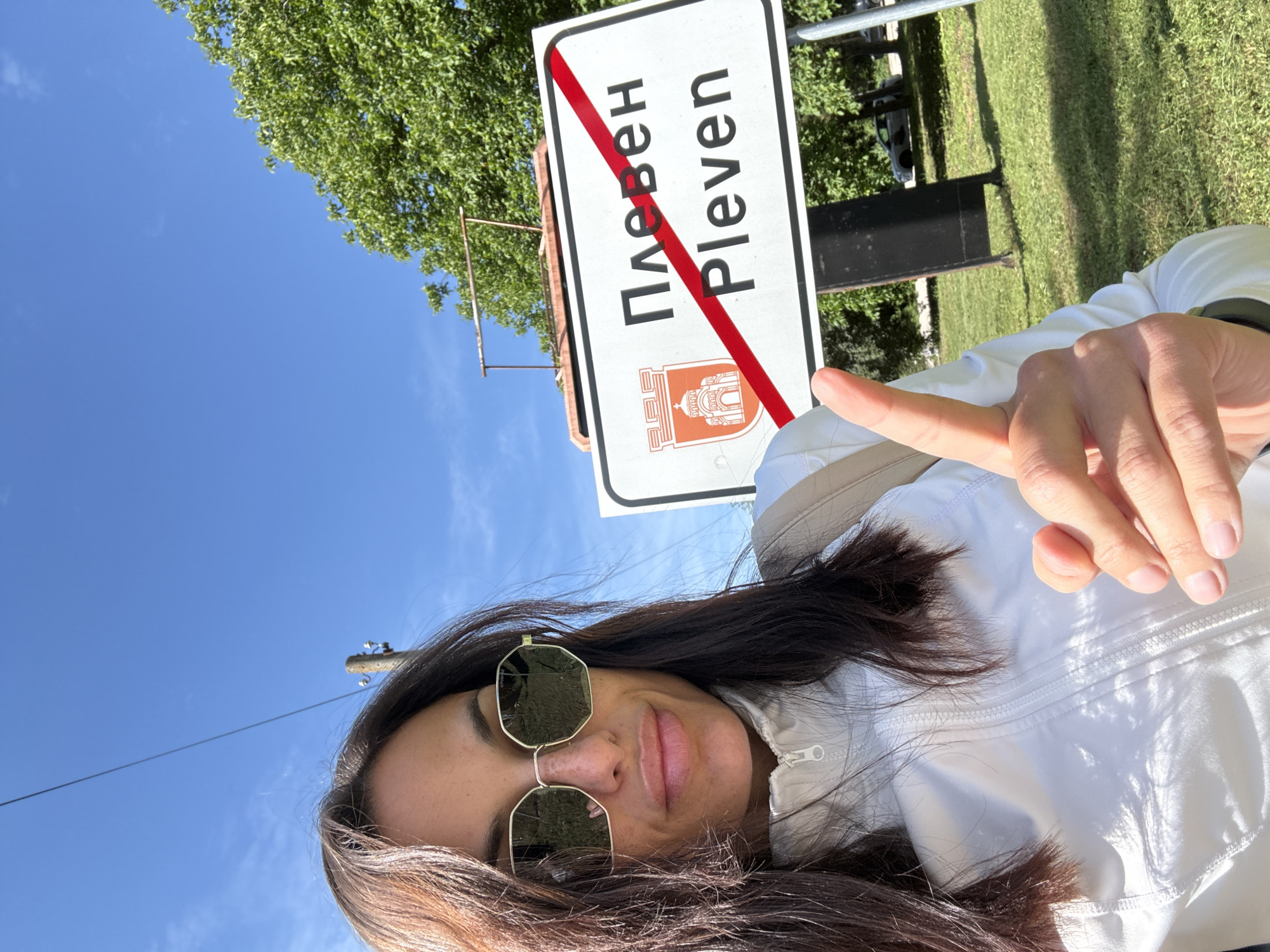 A medium shot captures a woman with dark hair, wearing sunglasses and a white zip-up jacket, posing for a selfie outdoors. She is in front of a road sign that reads "Плевен Pleven" with a red diagonal line crossing it, indicating the end of the city limits. A shield emblem is present on the left side of the sign. The background features a clear blue sky with some clouds, green trees, and a grassy area. A utility pole and wires are visible above the woman's head. The woman is pointing forward with her right index finger.