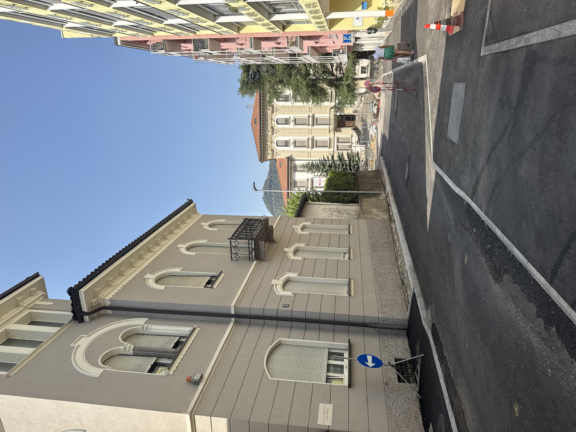The image depicts a street scene in Chiasso, Switzerland, under a clear blue sky. On the left, a grey, multi-story building with arched windows and decorative white trim is visible. A blue directional sign with a white arrow pointing left is placed on a stand in front of this building. The street in the foreground is paved with asphalt, with white lines marking lanes and a curb.
To the right, a row of colorful buildings, painted in shades of yellow, pink, and orange, lines the street. These buildings have balconies and numerous windows. Further down the street, a larger, ornate building with a red-tiled roof and cream-colored facade stands. Trees, including a tall evergreen, are present between the buildings.
In the mid-ground, a woman wearing a white top and green skirt walks away from the camera, carrying shopping bags. She is on the right side of the street, near a parking sign. Construction barriers and materials are visible further down the street, indicating ongoing work. A traffic cone with red and white stripes is placed on the right side of the road. The environment appears to be a mixed residential and commercial area.