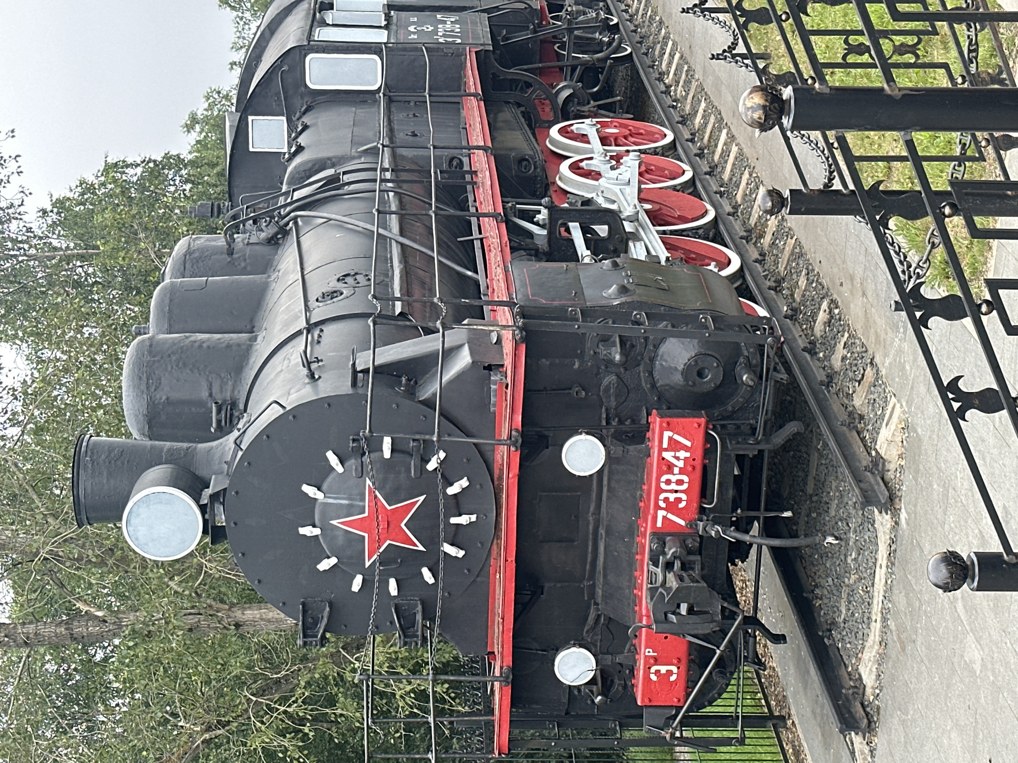 This is a full-shot, eye-level, outdoor photograph of a vintage black steam locomotive. The locomotive is positioned on a railway track and is the primary subject of the image. Its front features a prominent red star within a circle, and the number "738-47" is displayed on a red plate. The wheels are red with white rims. The locomotive is dark, with some red accents on its platform. Behind the locomotive, trees with green foliage are visible, suggesting an outdoor, possibly park-like setting. The sky is overcast. In the foreground, there is a black metal fence with ornate designs and chains. The locomotive is stationary on a set of tracks. Based on publicly available information, this steam locomotive is located in Petrozavodsk, Russia, specifically at the "Museum of Railway Technology" or a similar exhibit space. There are no people visible in the image.