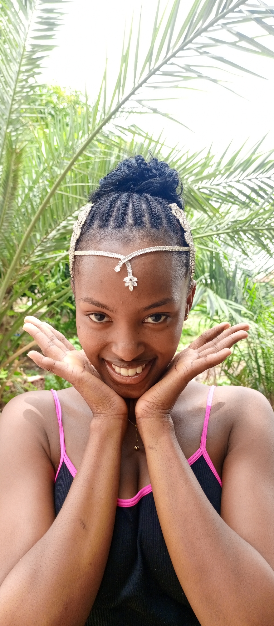 A young woman with dark skin is the central subject of this close-up, waist-up portrait. She looks directly at the viewer with a wide, joyful smile, revealing her teeth, and her eyes are bright and engaging. Her dark hair is styled in neat braids gathered into an updo or bun, and she wears a sparkling, silver-toned headpiece that drapes across her forehead and along her temples.
She is dressed in a black tank top or camisole with a vibrant pink trim visible around the neckline. A delicate gold chain necklace with a small, dark pendant hangs around her neck. Her hands are cupped playfully around her face, with her palms facing outward and fingers lightly touching her cheeks, enhancing her expressive pose.
The background is filled with lush, green foliage, specifically large, palm-like leaves, which are softly blurred, indicating a shallow depth of field. The lighting is bright and natural, suggesting the image was taken outdoors during the daytime. The setting appears tropical or subtropical, consistent with the provided location context of Keffi, Nigeria. The overall mood of the image is one of happiness and cheerfulness. There is no visible text.