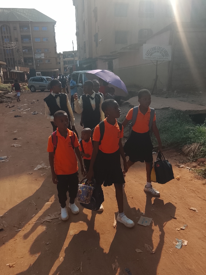 A group of schoolchildren, some in orange polo shirts and others in vests and bow ties, walk along a dusty road. They carry backpacks and bags. Buildings line the street, and a van and parked cars are visible in the background. A sign for "Exceeding Great Future Foundation and High School" is visible. The scene appears to be an everyday moment of children traveling to or from school.