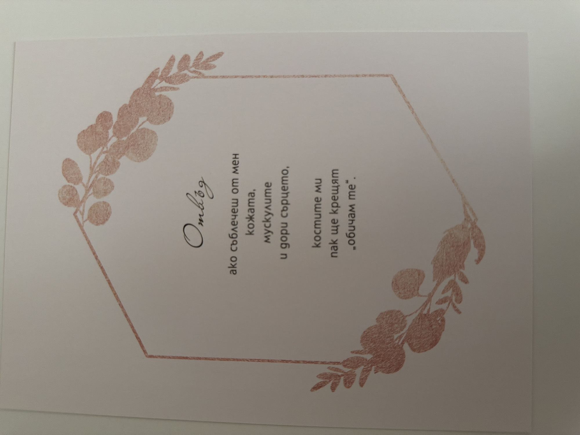 The image displays a white card with a poem written in Bulgarian script. The poem is centered within a geometric, hexagonal frame embellished with floral motifs in a rose gold color. The text is black and appears to be in a cursive font. Above the poem, the word "Отвъд" (Beyond) is written in a similar cursive style. The poem itself is a few lines long and discusses the skin, muscles, and heart. The card is placed on a plain surface, likely a table or desk. There are no people or animals visible in the image. The location of this object in the city of Pazardzhik, Bulgaria, cannot be determined from the image alone, as there are no contextual clues.