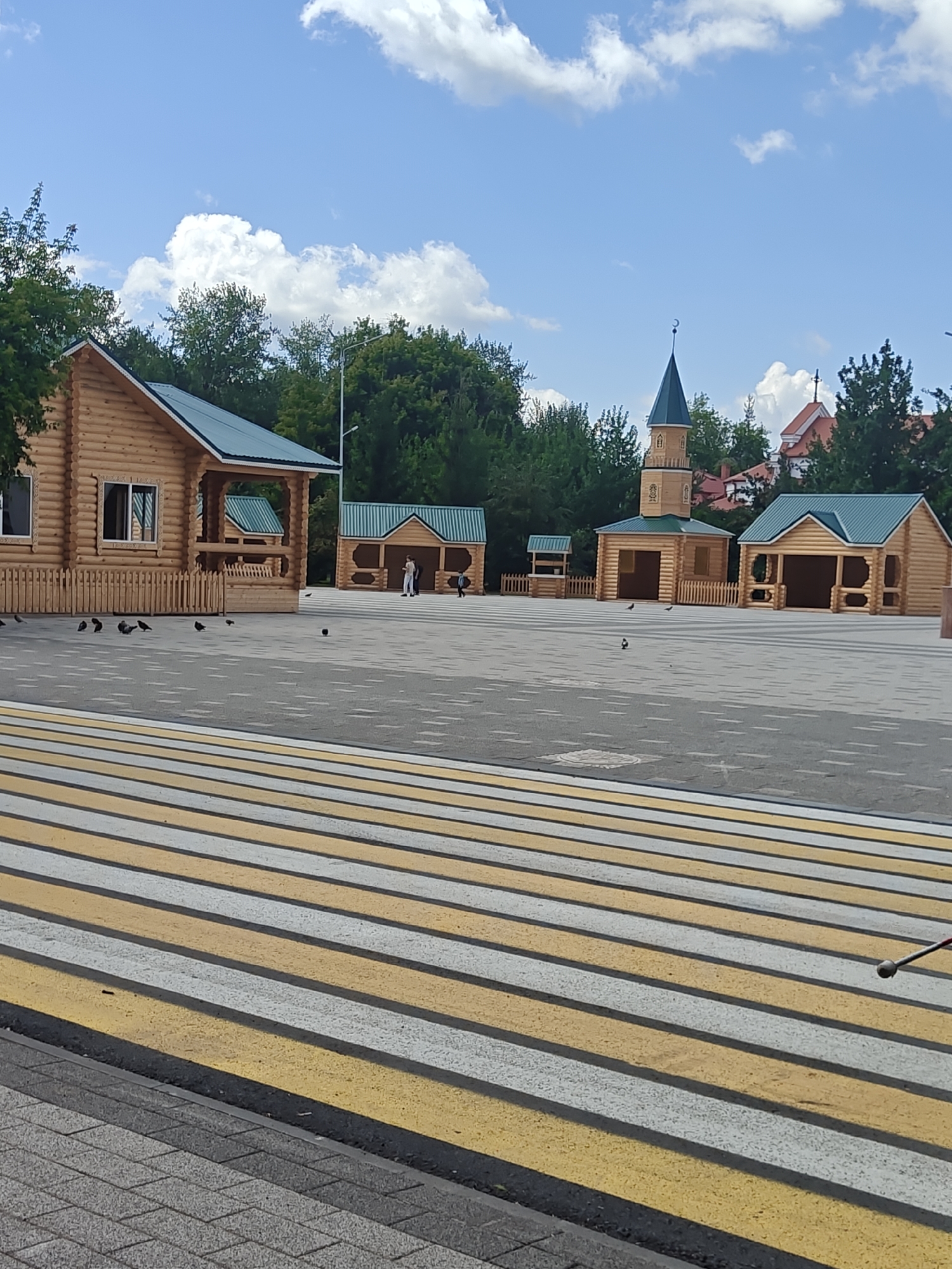 In Petropavl, Kazakhstan, this image captures a public area with several wooden structures, a prominent pedestrian crossing, and a clear blue sky with scattered clouds. The wooden structures appear to be small, rustic buildings, some resembling market stalls or small houses, with green roofs. One structure features a tall, slender tower topped with a crescent moon, characteristic of a minaret. The pedestrian crossing, painted with alternating yellow and white stripes, dominates the foreground, suggesting a street or public square. Pigeons are scattered across the paved ground. The overall environment is clean and open, with trees and other buildings visible in the background, indicating an urban or semi-urban setting.