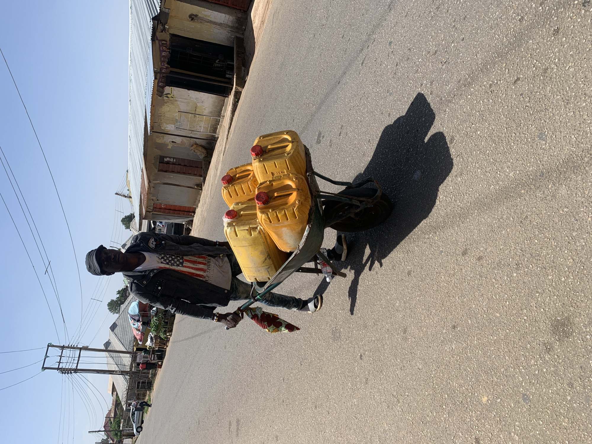 A man is pulling a green wheelbarrow on an asphalt road in Jos, Nigeria. The wheelbarrow contains four yellow jerrycans with red caps. The man is wearing a black cap, a dark denim jacket over a white t-shirt with a US flag design, and blue jeans. The road is bordered by buildings, including a corrugated metal structure. Power lines are visible in the background. The sky is clear and blue, indicating a sunny day. The man’s shadow is cast on the road.