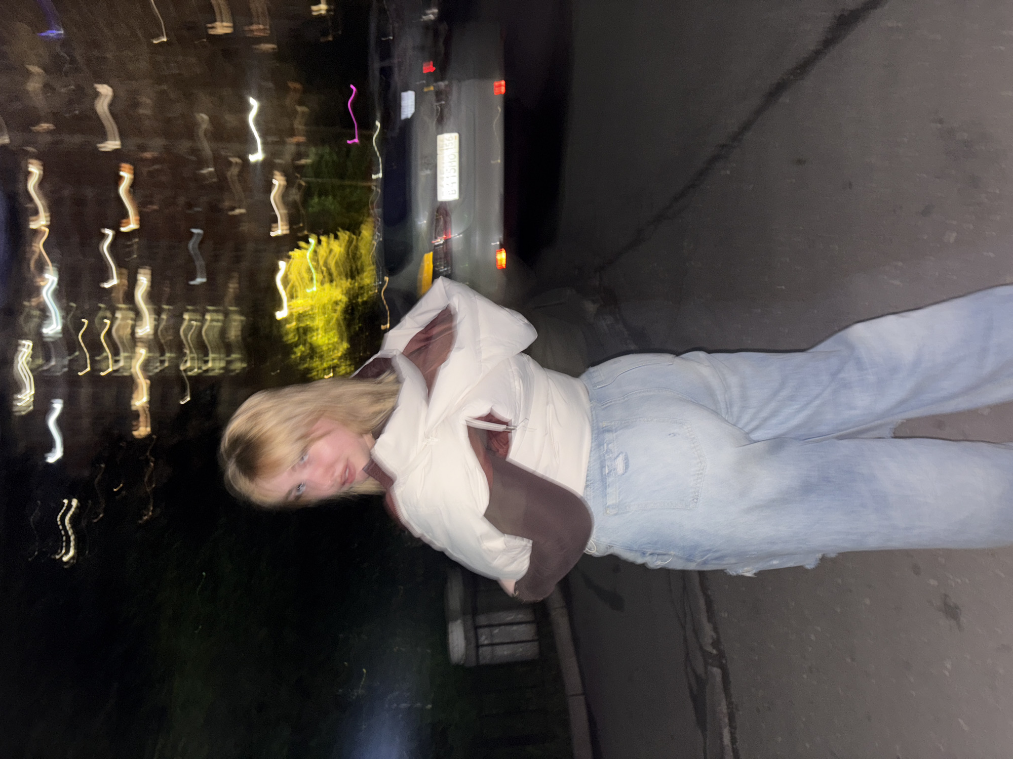 The image shows a young blonde woman standing in an urban environment at night. She is wearing a white puffer vest over a dark brown long-sleeved shirt and light blue ripped jeans. She is looking over her shoulder towards the camera. In the background, a light-colored car is parked on a street. The background also shows blurred lights and building facades, indicative of a city at night. The ground appears to be asphalt or pavement. The overall impression is that the photo was taken with a slight motion blur, common in low-light conditions. There is no specific visual information to confirm the location as Khimki, Russia.