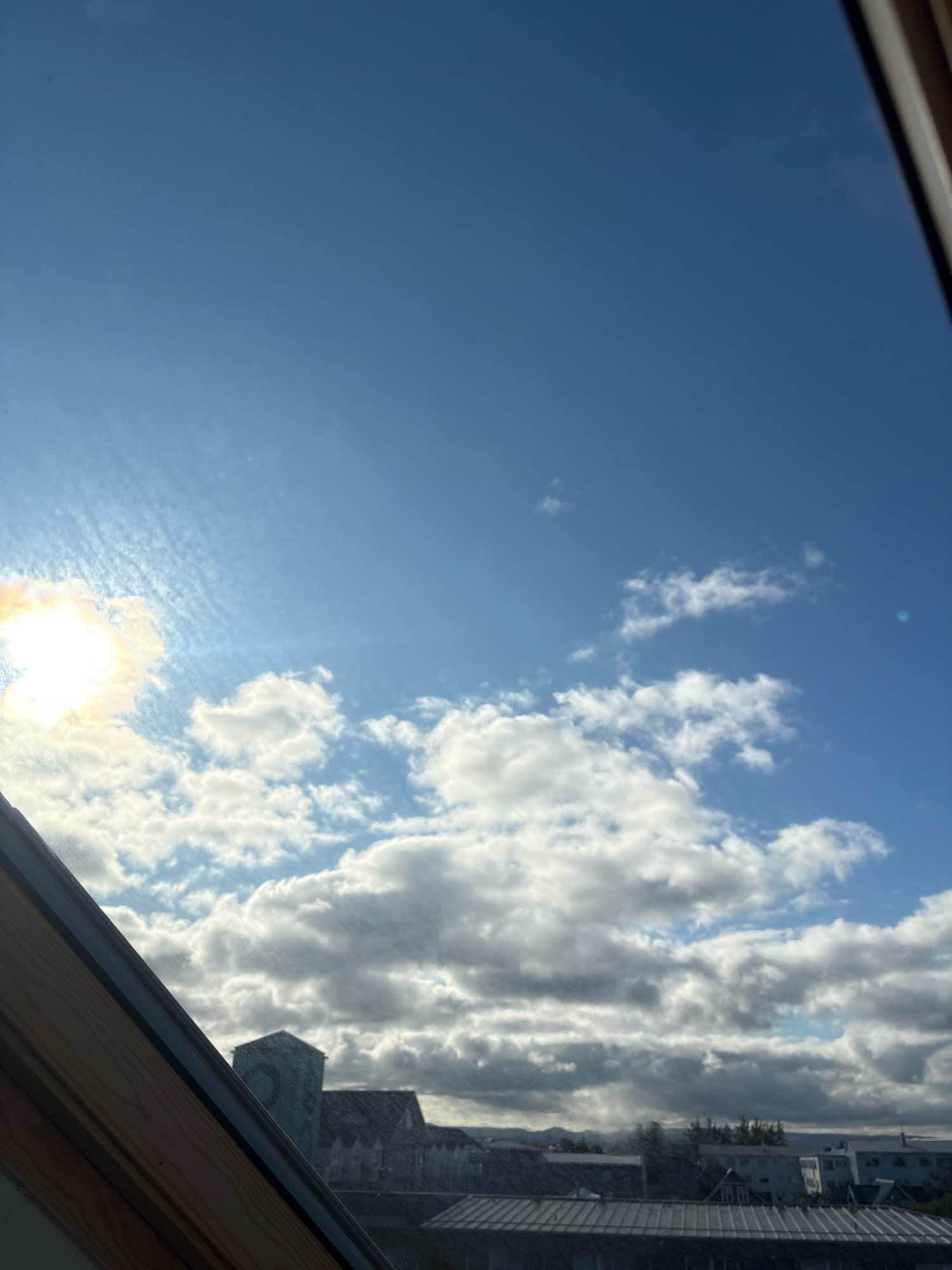 This is a vertical, eye-level, full shot of the sky and rooftops of Reykjavik, Iceland, as seen through a slanted window. The sky is a clear, bright blue with scattered white clouds, some of which are thin and wispy, while others are thicker and more dramatic. The sun is visible on the left side of the frame, a bright white orb with a yellowish halo, casting a slight glare. Below the sky, a line of buildings stretches across the horizon, their rooftops dark and indistinct against the bright sky. The buildings appear to be a mix of modern and older structures, with some featuring pitched roofs and others flat roofs. A prominent building with a green, angular roof is visible in the center-left of the frame. The window frame, made of wood, is visible in the foreground, running diagonally from the bottom left to the top right corner. The overall scene suggests a bright, possibly breezy day in Reykjavik.