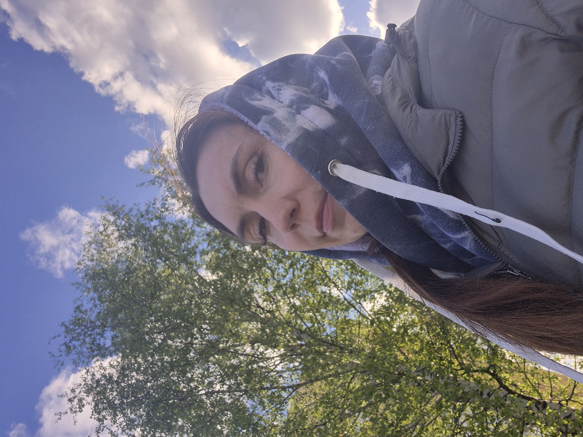 A selfie of a woman is taken in an outdoor setting. The background consists of a partly cloudy sky and treetops. The woman is wearing a grey jacket and a tie-dye hoodie with white drawstrings. The selfie appears to be taken while tilted. While I cannot confidently determine the exact location within Chaikovsky, Russia, the environment suggests a park or green space.