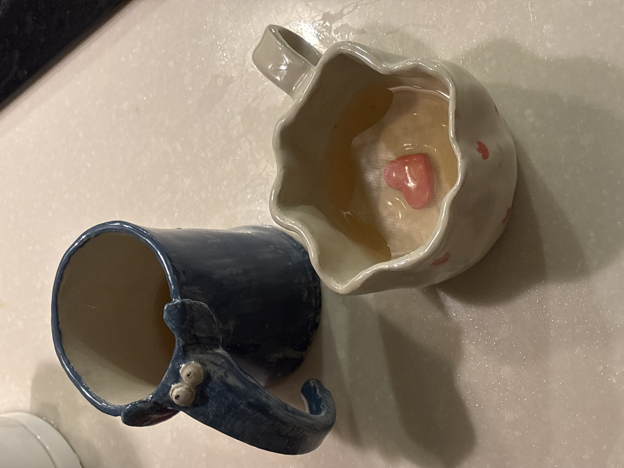 The image shows two ceramic mugs on a light-colored, speckled surface. One mug is light gray with a scalloped rim and a small pink heart at the bottom of the inside. It appears to contain a liquid. The other mug is blue with a whimsical handle featuring raised, rounded "eyes." The scene is not identifiable as Choeng Thale, Thailand, or any specific location. There are no people visible in the image.