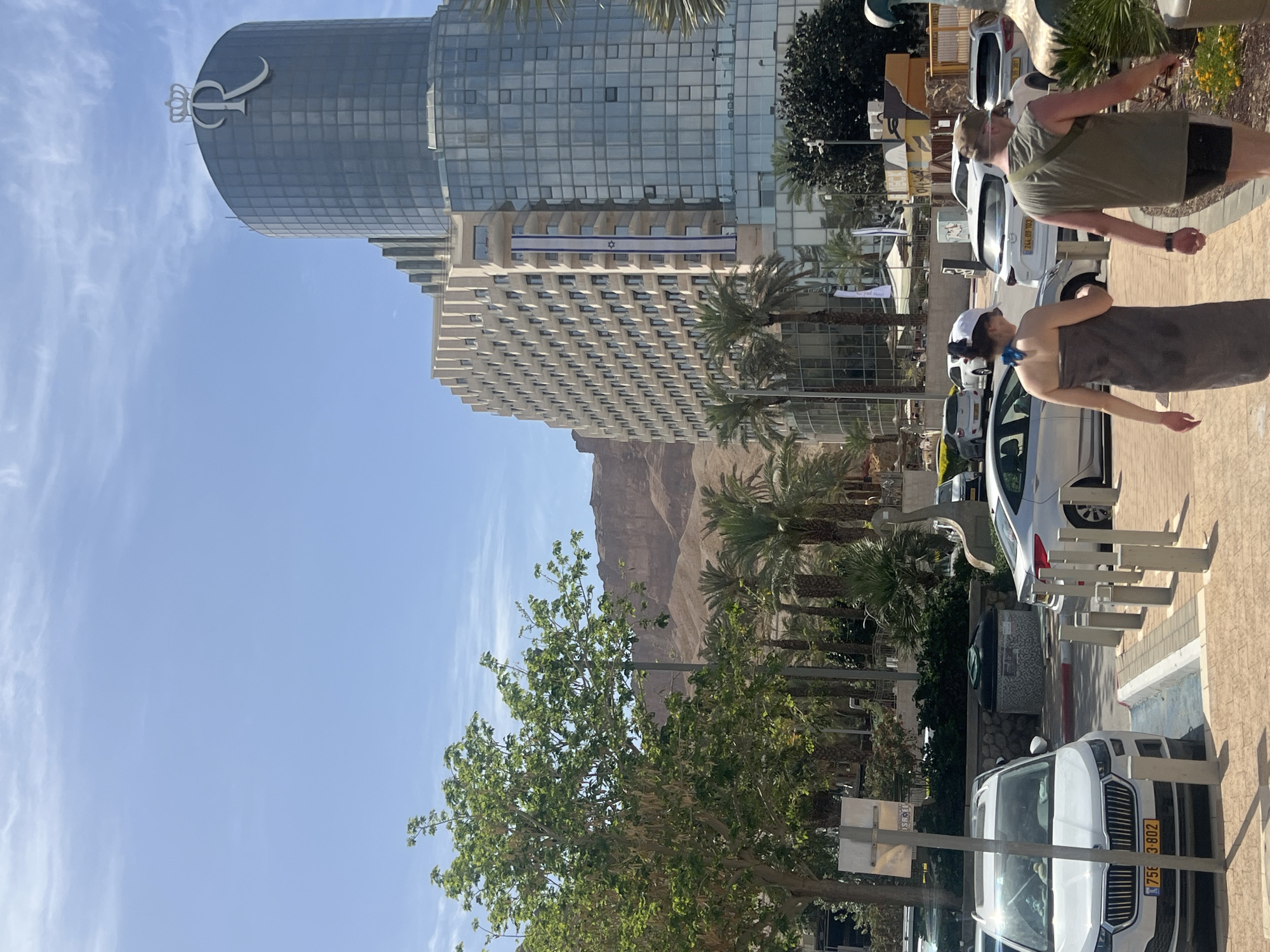 The image depicts a street scene in the city of Tamar Regional Council, Israel. In the foreground, two people are walking. One is wearing a towel. Cars are parked along the side of the street. A tall, modern hotel with a logo on top is visible in the background. Palm trees line the street. In the distance, mountains can be seen. The sky is clear and blue.