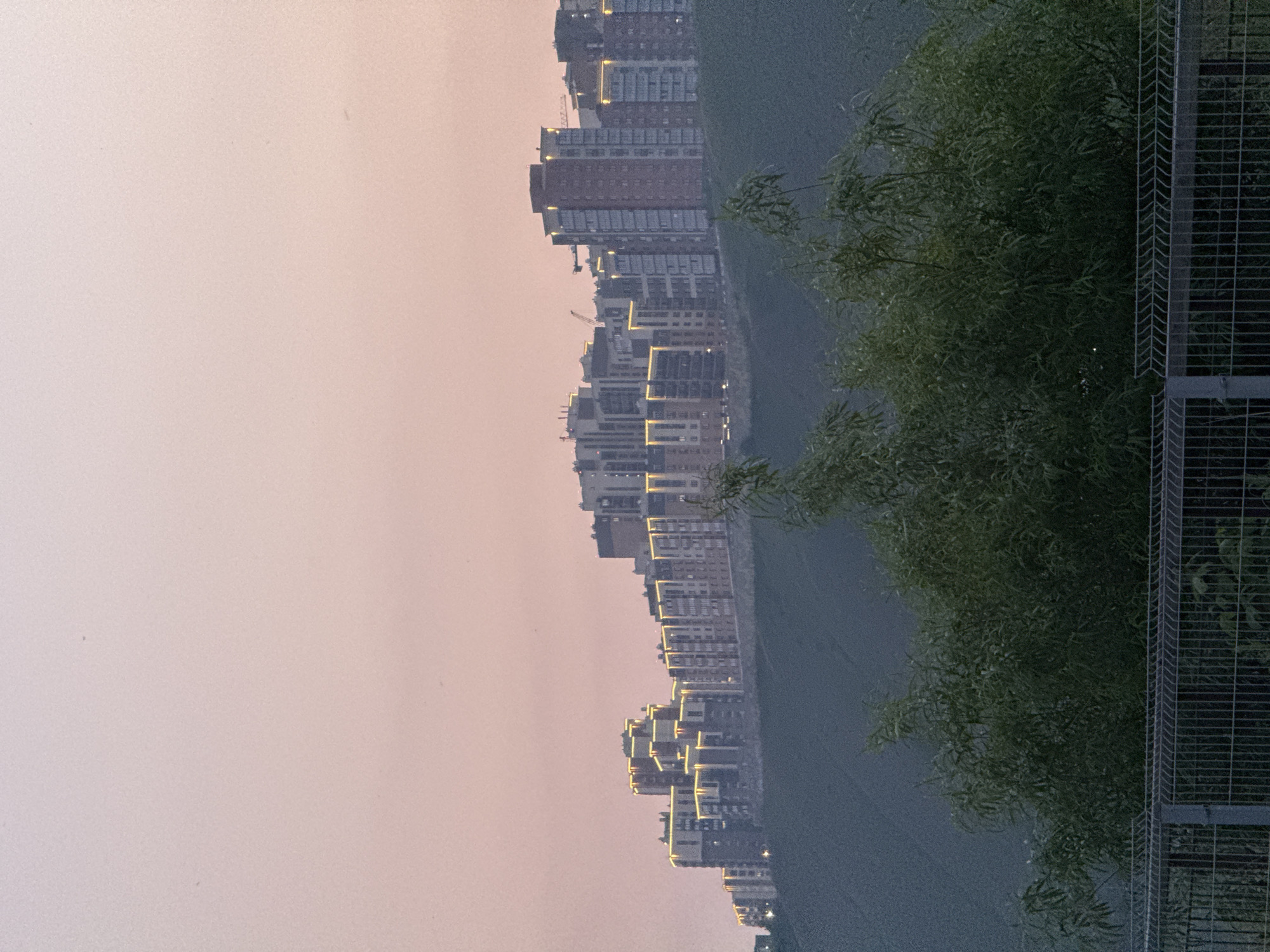 The image depicts a panoramic view of a modern residential complex situated on a hill overlooking a green, sloped terrain. The sky is a soft, hazy pink, suggesting either dawn or dusk. The buildings are a mix of architectural styles, with some featuring balconies and a prominent illuminated strip running along their upper edges, giving them a warm, inviting glow. In the foreground, the tops of lush green trees are visible, partially obscuring the view. Below the trees, a metal fence runs horizontally across the frame. The overall scene conveys a sense of urban development integrated with natural landscape. This residential complex is likely located in the area of the Yenisei River, near the city of Krasnoyarsk, Russia.