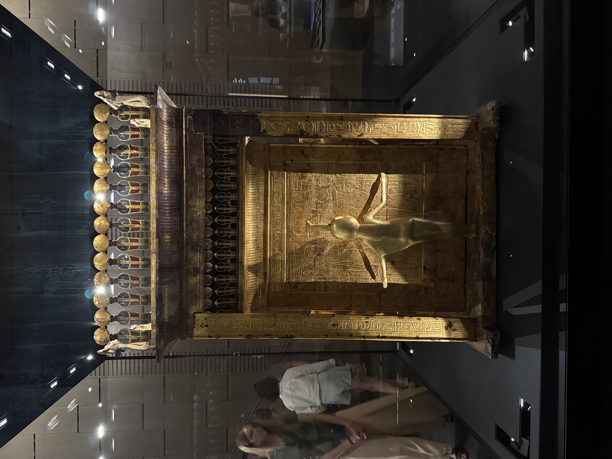 In Kafrat Nassar, Egypt, a golden sarcophagus is displayed in a museum. The sarcophagus is ornate, with hieroglyphs and figures adorning its surface. It stands within a glass case, illuminated by spotlights. Two women are visible in the background, looking at the exhibit. The museum environment is modern, with clean lines and reflective surfaces.