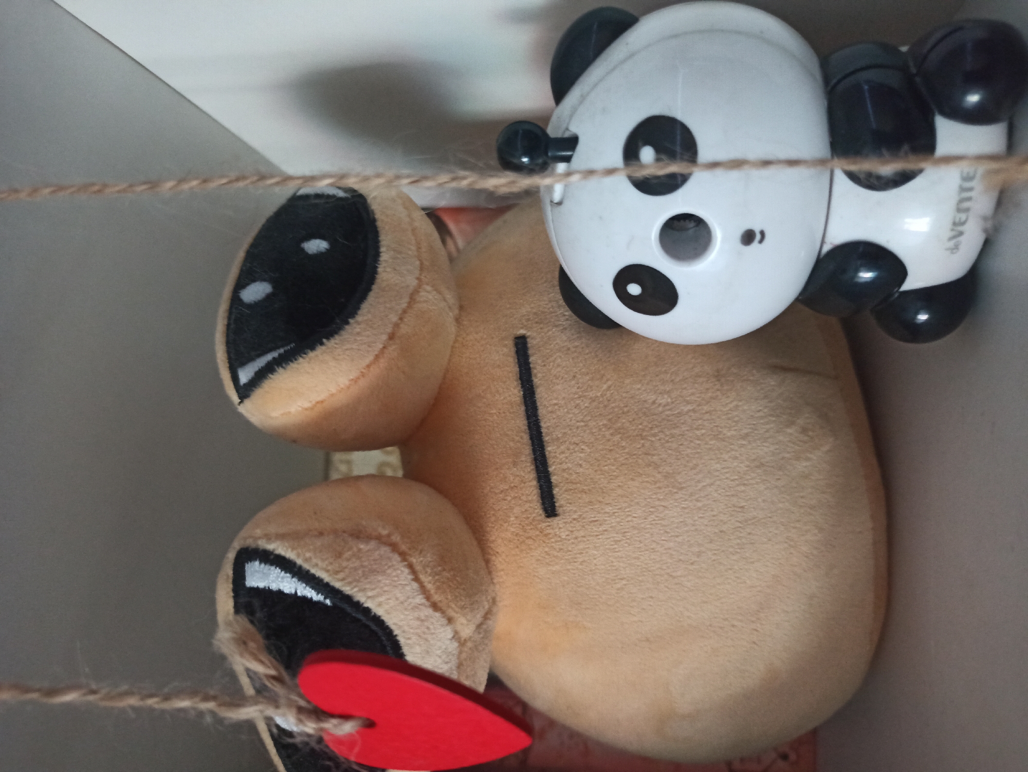 The image displays a close-up view of two stuffed toys, likely on a shelf. On the left is a large, light brown, vaguely animal-shaped plush toy with two large black eyes outlined in white, and a small red heart hanging from a string on its left side. A thin black horizontal line is visible on its body. To the right of this larger toy is a smaller, white and black stuffed panda bear. The panda has black ears, black eye patches, and a black mouth. It appears to be a pencil sharpener, with a circular opening on its forehead. A piece of twine runs vertically through the center of the image, obscuring part of the panda. The background is a pale, greyish surface, possibly a wall or the interior of a shelf. The lighting suggests an indoor environment. There is no visible indication of the city of Promyshlennaya, Russia, within the image itself.
