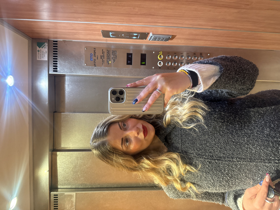 An individual, female, is depicted taking a selfie within an elevator in Paris, France. She possesses long, wavy blonde hair with visible darker roots, parted down the middle, and wears dark red lipstick. Her attire consists of a dark grey, textured, possibly boucle-knit jacket over a light-colored, ribbed long-sleeved garment. Her left hand, extended to hold a light-colored smartphone with a ribbed case, shows dark blue glitter-tipped fingernails and a ring on her ring finger. Her right wrist is adorned with multiple bracelets, including a yellow beaded one and a dark elastic band. In her right hand, positioned lower in the frame, she holds a set of keys, including a car remote fob, and her fingernails exhibit a different multi-colored polish. The elevator interior features reflective metallic doors on the left and a wooden panel on the right. Overhead, two circular light fixtures are visible, one emitting a strong glare. A sticker on the metallic panel displaying the mirrored French text "CONTROLE TECHNIQUE ASCENSEURS" confirms the location in France.