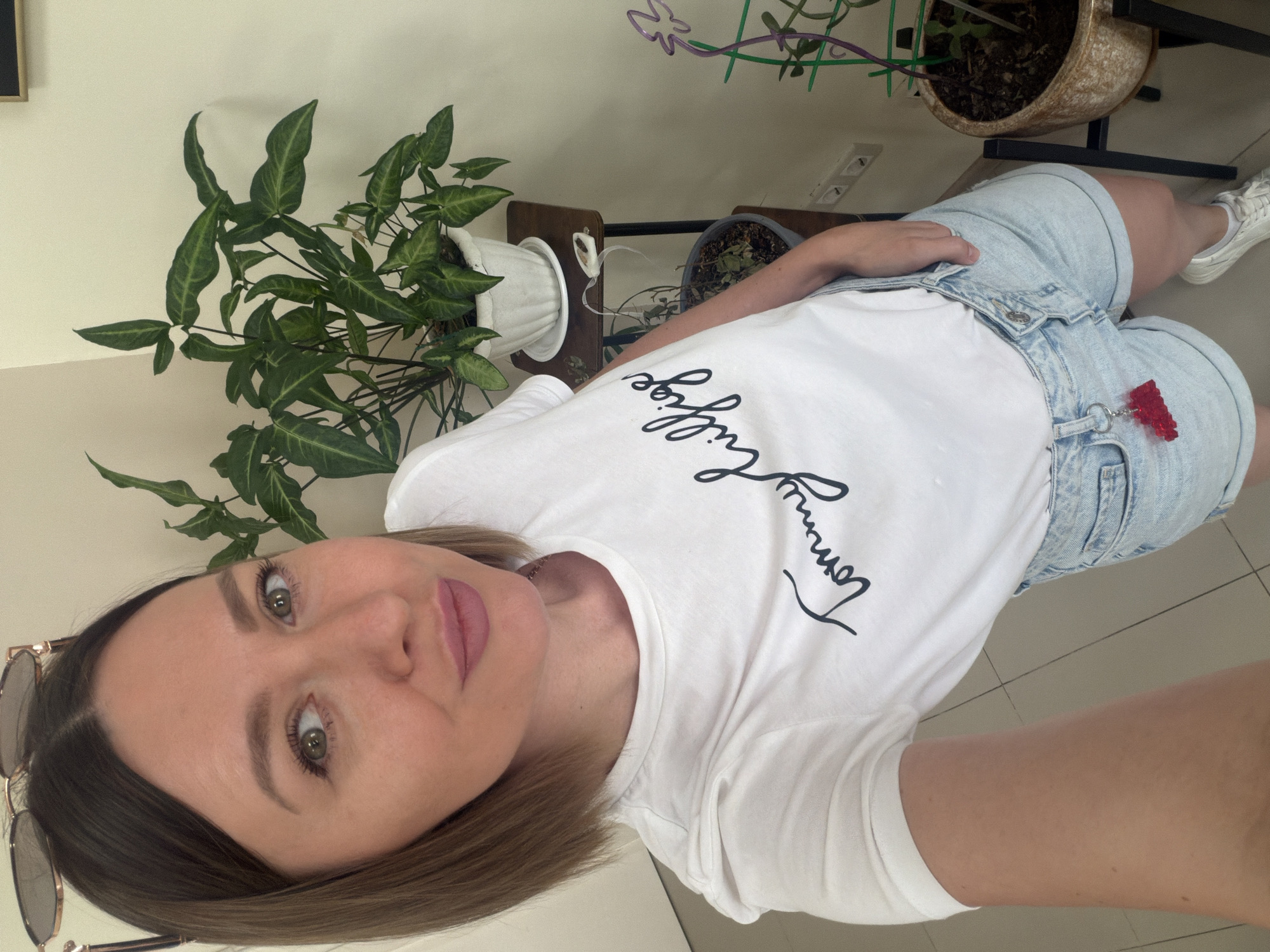 The image shows a woman taking a selfie indoors. She is positioned slightly off-center, looking directly at the camera with a neutral expression and a faint smile. She has shoulder-length brown hair, brown eyes, and light makeup. She is wearing a white short-sleeved t-shirt, light blue denim shorts, and white sneakers. A pair of sunglasses with a metallic frame is hooked onto the neckline of her t-shirt. A red, translucent gummy bear-shaped keychain is attached to a belt loop on her shorts.
The setting appears to be a well-lit indoor space, likely a home, with a light-colored wall and a tiled floor. To the left of the woman is a large potted plant with green, arrow-shaped leaves, placed in a white ribbed pot. Behind her, a wooden stand holds another plant pot with dark soil, and a third plant pot is visible further back, containing a plant with some bare branches and green leaves, possibly with a small trellis. An electrical outlet is visible on the wall behind the plants. The overall impression is casual and relaxed, captured during the daytime.
Visible text on her t-shirt reads "Tommy Hilfiger" in a black cursive script. Based on the provided context, the location is Rostov-on-Don, Russia.
