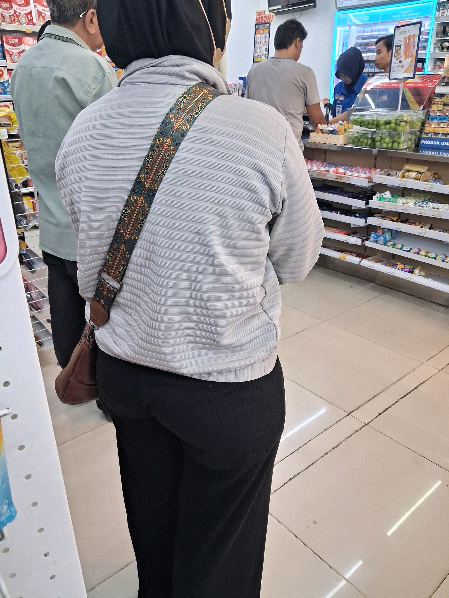 A rear view shows three individuals inside a bright, tiled convenience store in Surakarta, Indonesia. The foreground features a person from behind, wearing a light gray, horizontally ribbed long-sleeved shirt with a dark, patterned strap draped across their back, a black hijab, and black pants. A brown shoulder bag is visible on their hip. To the left, another person stands wearing a light green long-sleeved shirt and black pants. In the background, two individuals, one in a gray t-shirt and the other in a dark blue uniform with a hijab, are interacting at a counter with shelves of products behind them. The shelves display various packaged goods and some fresh produce. The overall environment suggests a typical retail setting.