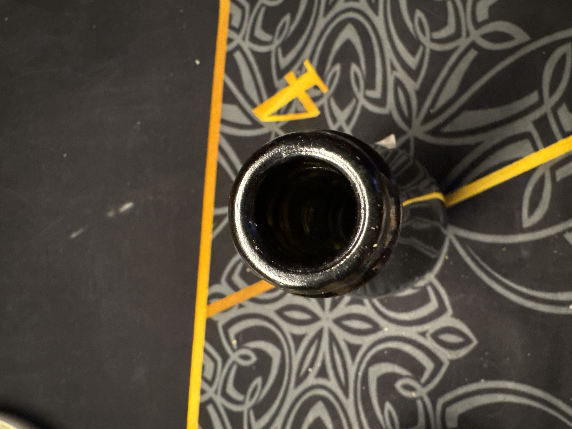 A high-angle close-up shows the open mouth of a dark-colored bottle. The bottle sits on a dark surface with a stylized pattern of light gray, possibly floral or Celtic knot designs. A thin yellow line intersects the pattern near the bottle. In the upper right of the frame, part of another yellow object is visible near the patterned surface. The location in the city of Khimki, Russia cannot be determined from the provided image.