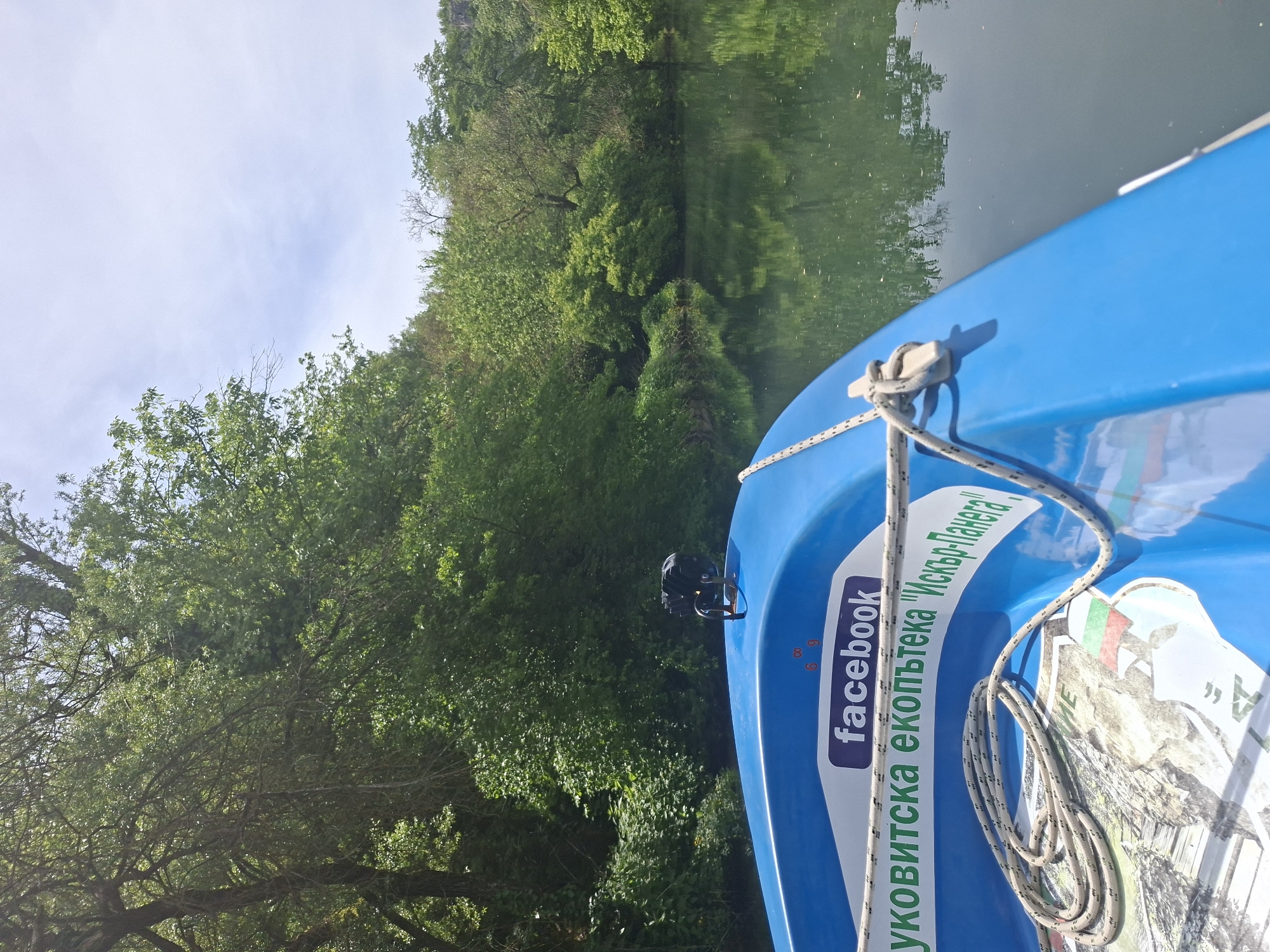 The image shows the bow of a bright blue boat on a body of water with green foliage and trees visible in the background. The water's surface reflects the surrounding trees and sky. A rope is tied to the boat's cleat. A sticker on the boat features the Facebook logo and Bulgarian text. Based on the text on the sticker and likely geographic location, the image is potentially taken in the region of Petrevene, Bulgaria.
