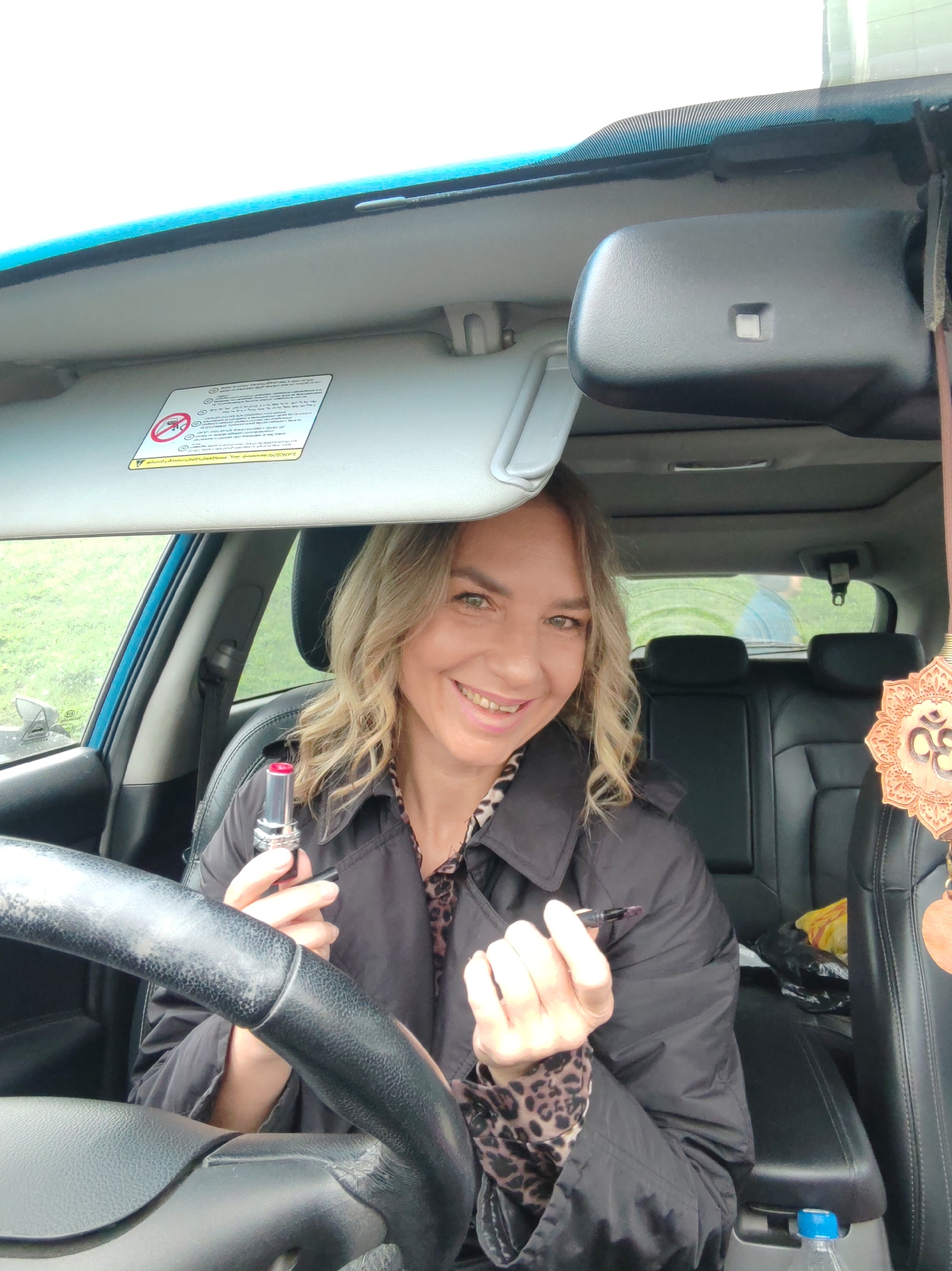 A smiling woman with wavy blonde hair is seated in the driver's seat of a car, looking directly at the camera. She wears a black trench coat over a leopard print top. In her left hand, she holds an open silver-cased lipstick with a bright red/pink shade, and in her right hand, she holds a thin black pen-like object, possibly an eyeliner or lip liner. Her expression is cheerful and relaxed.
The setting is inside a car, with the steering wheel partially visible in the foreground. The car's interior features black seats and a raised sun visor. Through the windows, a green, natural outdoor background is visible, suggesting daytime. A rearview mirror is in the upper right. Hanging from the rearview mirror area is a decorative wooden pendant with the 'Om' symbol carved into it. A blue-capped water bottle is partially visible in the center console area.
On the sun visor, a white warning label with black text and symbols is prominent. The text, in Cyrillic script, includes "ВНИМАНИЕ" (Attention) and "Опасность! Не устанавливайте детское удерживающее устройство..." (Danger! Do not install child restraint device...). A pictogram showing a disallowed rear-facing child seat is also visible, indicating warnings related to airbags.