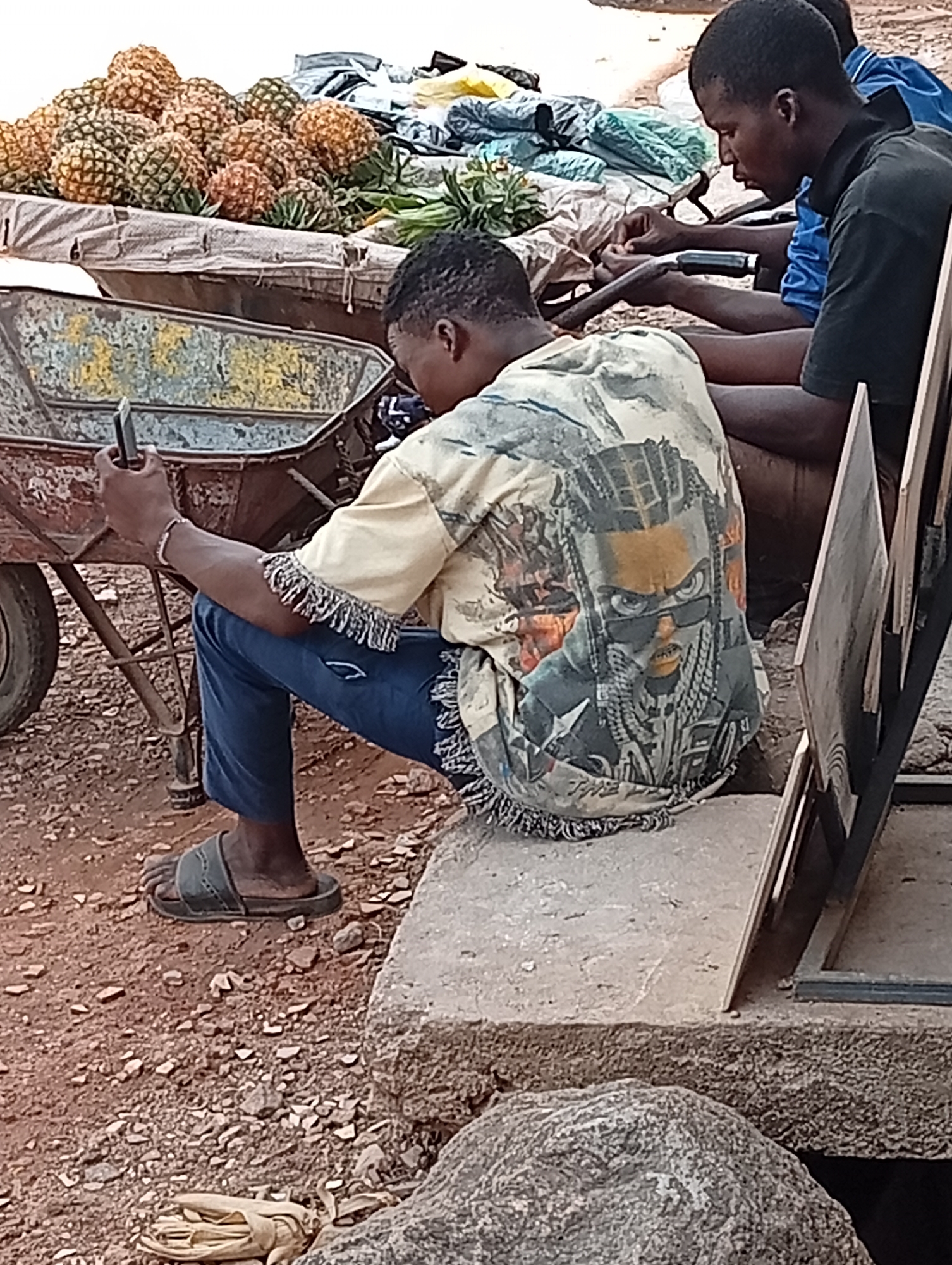 The scene is in Maiburiji, Nigeria. A man sits on a concrete block, facing away from the camera, holding a phone in his left hand. He wears a fringed, patterned shirt with a graphic image of a person wearing sunglasses, blue jeans, and slides. To his left, a rusted wheelbarrow holds a pile of pineapples. Beside him to the right, another man sits on a bench, facing right, with another man partially visible behind him. The ground is red earth with scattered stones. A tied bundle of straw lies near the base of the concrete block.