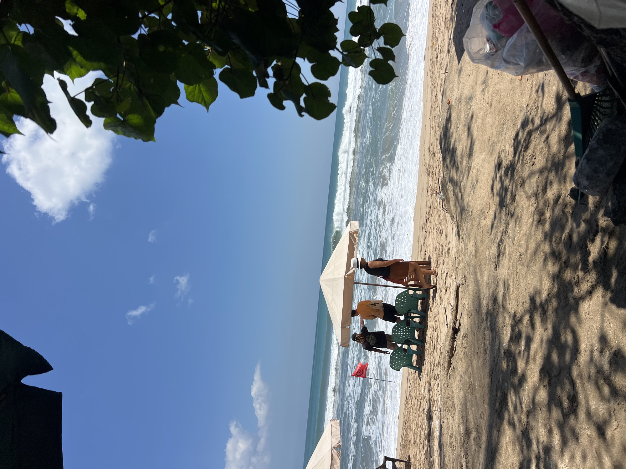 The image depicts a sunny day at Kuta Beach, Indonesia, with a vibrant blue sky and scattered clouds. In the foreground, the sandy beach is partially shaded by foliage, casting dappled shadows. A rake and a trash bag filled with collected debris are visible. Further down the beach, closer to the ocean, are several green plastic chairs and a beige beach umbrella. Three people are present near the chairs and umbrella: one person wearing a black t-shirt and hat, another in an orange t-shirt, and a third person in a black top and brown skirt. A red flag with a circular symbol is planted in the sand. The ocean is active, with moderate waves breaking on the shore. In the distance, another beach umbrella is visible, and faint outlines of surfers can be seen on the water.