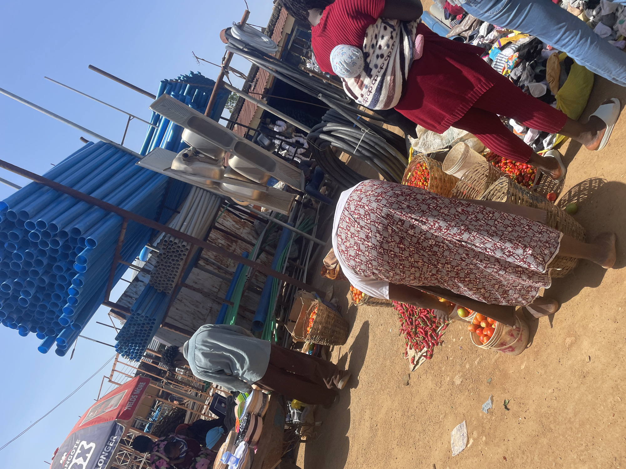This high-angle, tilted photograph captures a bustling, sunlit open-air market scene in Gura Top, Nigeria. The bright, clear blue sky indicates fair weather, and strong shadows suggest it is midday or early afternoon.
The market is a vibrant mix of goods and people. In the midground, a woman dressed in a red ribbed outfit carries a baby secured in a patterned cloth on her back. She stands near several woven baskets filled with bright red peppers and ripe tomatoes. To her right, another woman, wearing a white top and a patterned red and white skirt, is bent over, tending to more produce displayed in baskets and on the ground. A man in a light blue/grey shirt and dark trousers is visible to the left, facing away from the camera, near other market items. Part of a person's legs in blue jeans can be seen at the top right, standing over a heap of clothing or fabric.
The market stalls offer a diverse range of products. Prominently featured on the left are numerous bundles of bright blue PVC pipes stacked on metal frameworks, alongside several silver-colored metal sinks or washbasins. Coiled black hoses or cables are also visible on racks. The ground beneath the vendors is dirt, with occasional pieces of discarded plastic litter. In the background, a market umbrella or canopy with red and blue sections displays text that reads "ANGOTE 3X LONGER" and "FIELDS MORE," indicating commercial branding. The overall impression is a busy, functional marketplace where a wide array of necessities are bought and sold.