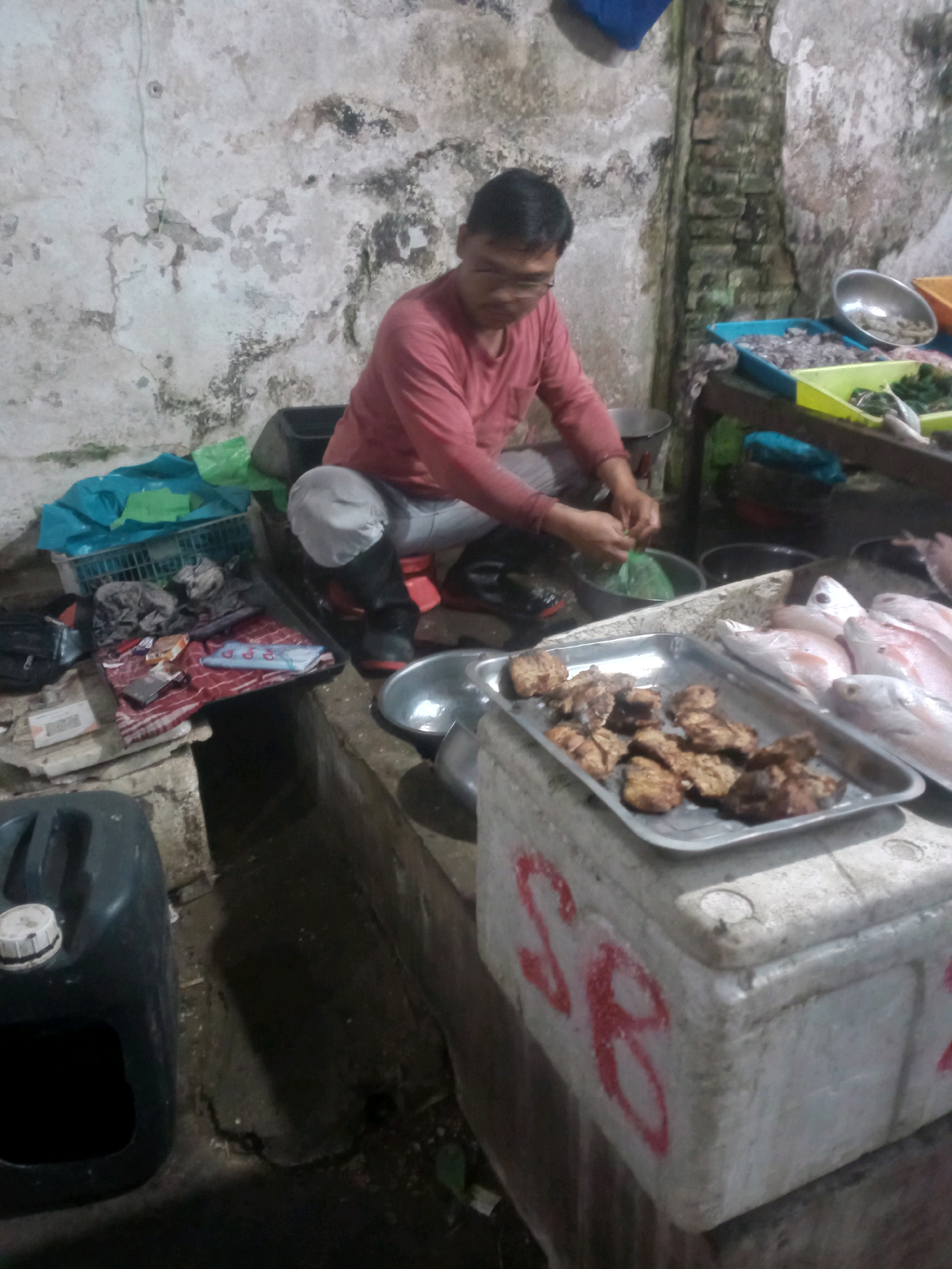A man, likely a vendor, is crouched down in front of a stall in what appears to be an outdoor market in Maguwoharjo, Indonesia. The setting is rustic, with a cracked and weathered concrete wall as the backdrop and a brick pillar visible on the right. The ground is uneven and appears to be wet or damp.
The man is wearing a long-sleeved pink shirt, light grey pants, and dark boots. He is actively engaged in his work, as evidenced by his hands holding a green plastic bag and seemingly preparing something. He is wearing glasses.
In front of him, on a counter or table, are various items related to selling food. A silver tray holds several pieces of cooked, browned food items, possibly meat or seafood. Next to this tray, another silver tray contains several whole fish, appearing to be of the same type, with pinkish-white flesh. There are also several containers and baskets, some holding what looks like raw ingredients or prepared food. A large, dark plastic container with a white cap is positioned on the lower left of the frame.
The overall impression is a scene of commerce and preparation within a local marketplace. The lighting is somewhat dim, suggesting it might be early morning, late evening, or an indoor/covered area with limited light.