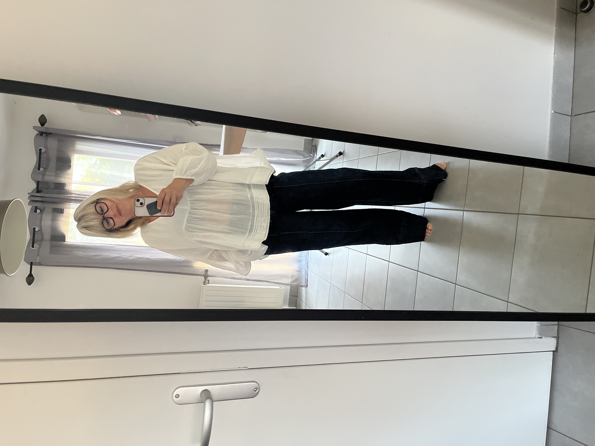 A woman with blonde hair and glasses is taking a mirror selfie in what appears to be a home. She is wearing a loose-fitting white blouse and dark wide-leg pants. Her phone is visible in her hand. The background shows a tiled floor, a radiator, sheer curtains, and a glimpse of the outdoors through a window. A door with a silver handle is visible on the left side of the frame. The scene is well-lit, suggesting it is daytime. There is no indication of the city of Chambœuf, France, within the image itself.
