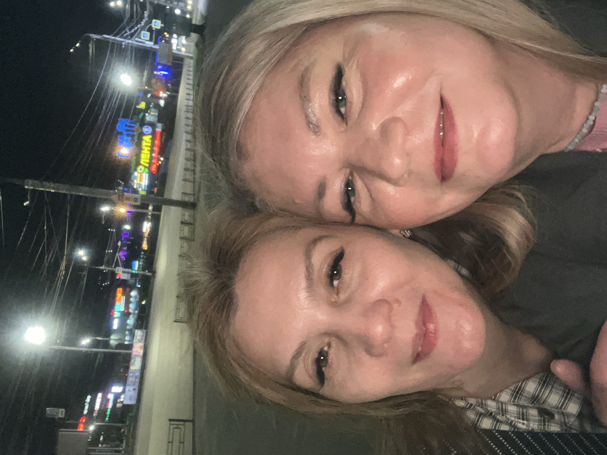 Two light-skinned women are shown in a close-up selfie taken at night. The woman at the top of the frame has blonde hair and blue eyes. The woman below has brown hair and eyes. They are close, with their heads nearly touching. In the background, there is an out-of-focus view of a city street at night, lit by streetlights and neon signs. One sign distinctly reads "ЛЕНТА," which transliterates to "LENTA," a Russian retail chain. The location is likely Izhevsk, Russia, where there is a LENTA store.