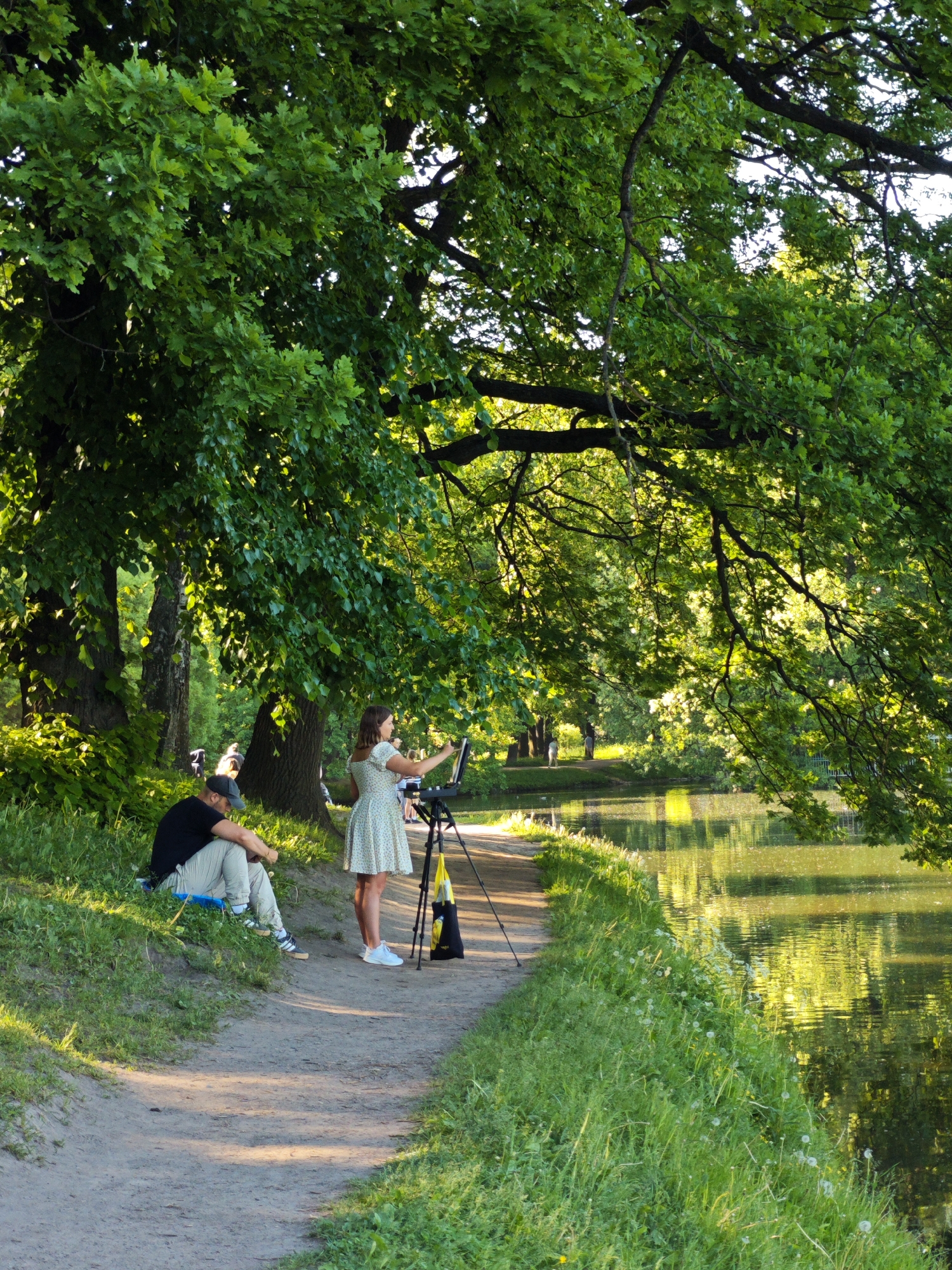This outdoor image depicts a park scene in Saint Petersburg, Russia, under bright, dappled sunlight. A woman stands on a dirt path facing right, wearing a light-colored, patterned short-sleeved dress and white sneakers. She is interacting with a black tripod that supports a flat panel or canvas, her right arm raised. A yellow and black bag hangs from the tripod at its base. To her immediate left, a man is seated on a small blue mat on the grassy bank, facing away from the viewer, wearing a black t-shirt, light-colored pants, and a baseball cap. His head is lowered.
The dirt path extends from the lower center into the background, running parallel to a body of water, likely a pond or lake, on the right. The water's surface reflects bright, yellowish-green light and the surrounding foliage. Dense green leaves, primarily from large trees resembling oaks, dominate the upper portion of the frame and cast shadows across the scene. The grassy bank adjacent to the path features tall green grass and scattered white wildflowers. Several indistinct figures are visible in the background, further along the path and across the water.