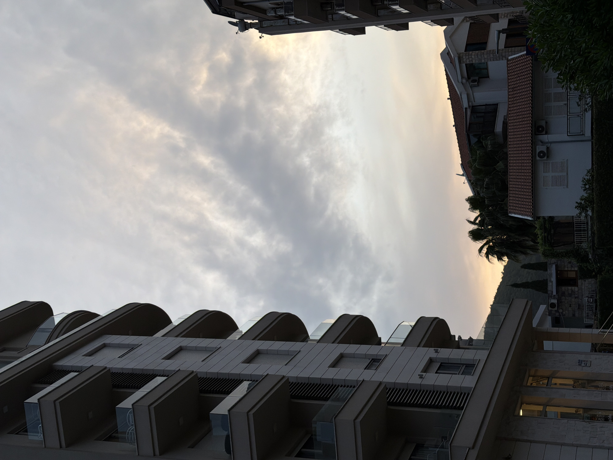 A low-angle shot shows the sky and buildings in Budva, Montenegro. The sky is filled with gray and white clouds, with a hint of golden light towards the right side of the frame. Buildings frame the sky. At the bottom is a modern building with multiple protruding balconies. The building is gray and has a contemporary design. Another building is partially visible on the right side of the frame. It has a red-tiled roof and white walls. There are green trees and a palm tree beside the building. A third building is at the top of the image with a grayish color and a satellite dish. The overall perspective is looking upwards, giving prominence to the sky and the architectural elements of the buildings.
