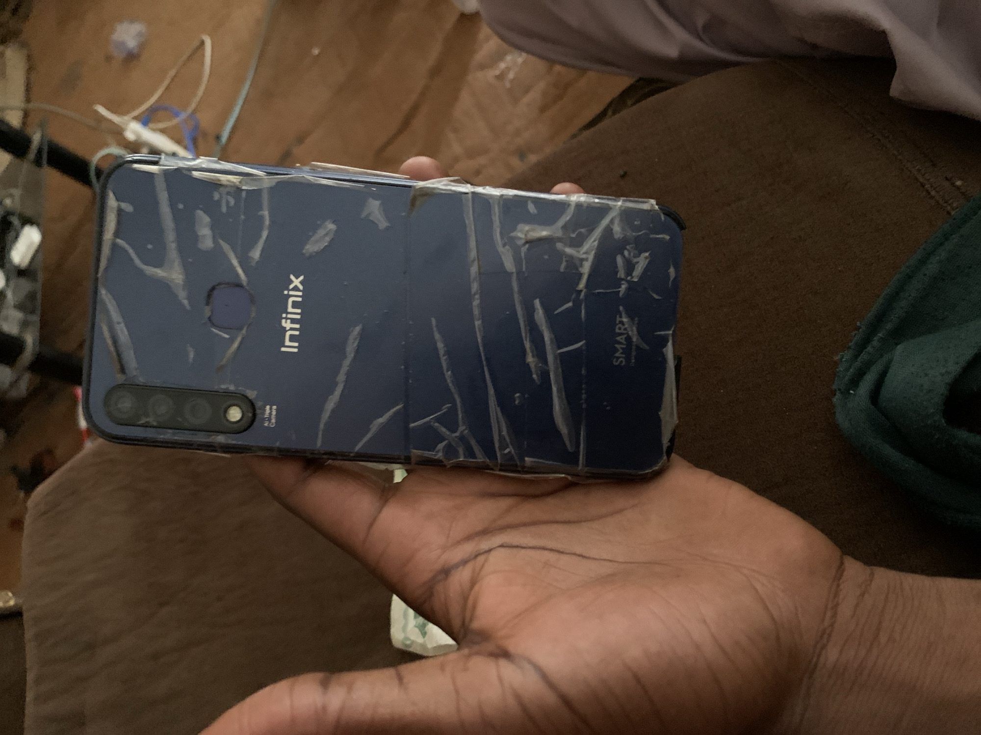 A dark blue Infinix brand smartphone is held by a person's hand, obscuring the lower left portion of the device. The phone's back is covered in what appears to be a cracked plastic protective film, with additional tape applied to secure parts of it. A triple camera setup is visible on the upper left of the phone's rear, labeled "AI Triple Camera." The Infinix logo is centered below the cameras, and the word "SMART" is visible near the bottom. The background is out of focus, showing a brown upholstered surface, possibly a couch, and some electronic equipment and cables on a wooden surface. The image's lighting suggests an indoor setting. The image was taken in Jos, Nigeria.