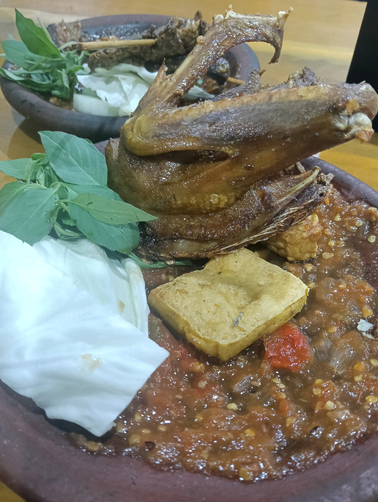 In a close-up, high-angle shot, a plate of Indonesian food is presented on a wooden table. The dish features a serving of fried duck or chicken, with prominent wings, seasoned with spices. Alongside the poultry, there is a dollop of sambal, a spicy chili paste, and pieces of fried tofu. Fresh basil leaves and a piece of white cabbage are also visible. The food is served in a traditional clay mortar. The overall setting suggests a casual dining experience in Ardirejo, Indonesia.