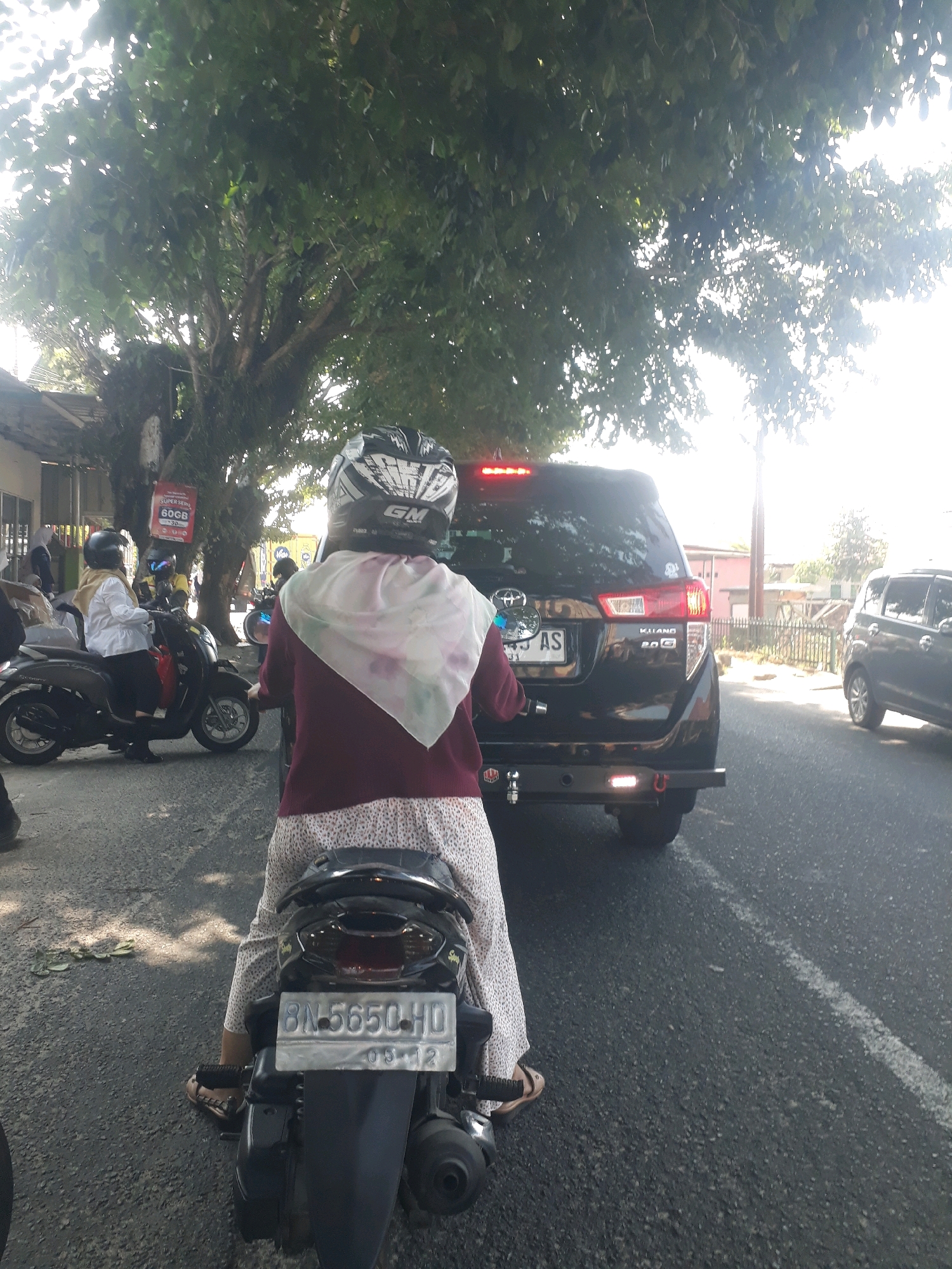 The image depicts a street scene in a city. In the foreground, a person wearing a helmet and a maroon shirt over a patterned white skirt rides a scooter. Their feet, clad in brown sandals, are visible. The scooter has a license plate that reads "BN-5650-HD" and "09-12". To the right of the scooter, a black SUV is partially visible, displaying a license plate with characters including "AS". Another black car is parked further down the street on the right. In the background, a large tree with dense green foliage casts shadows on the road. Buildings and a street light are also visible in the distance. The sky is bright, suggesting daylight. The overall scene is of traffic on a street.