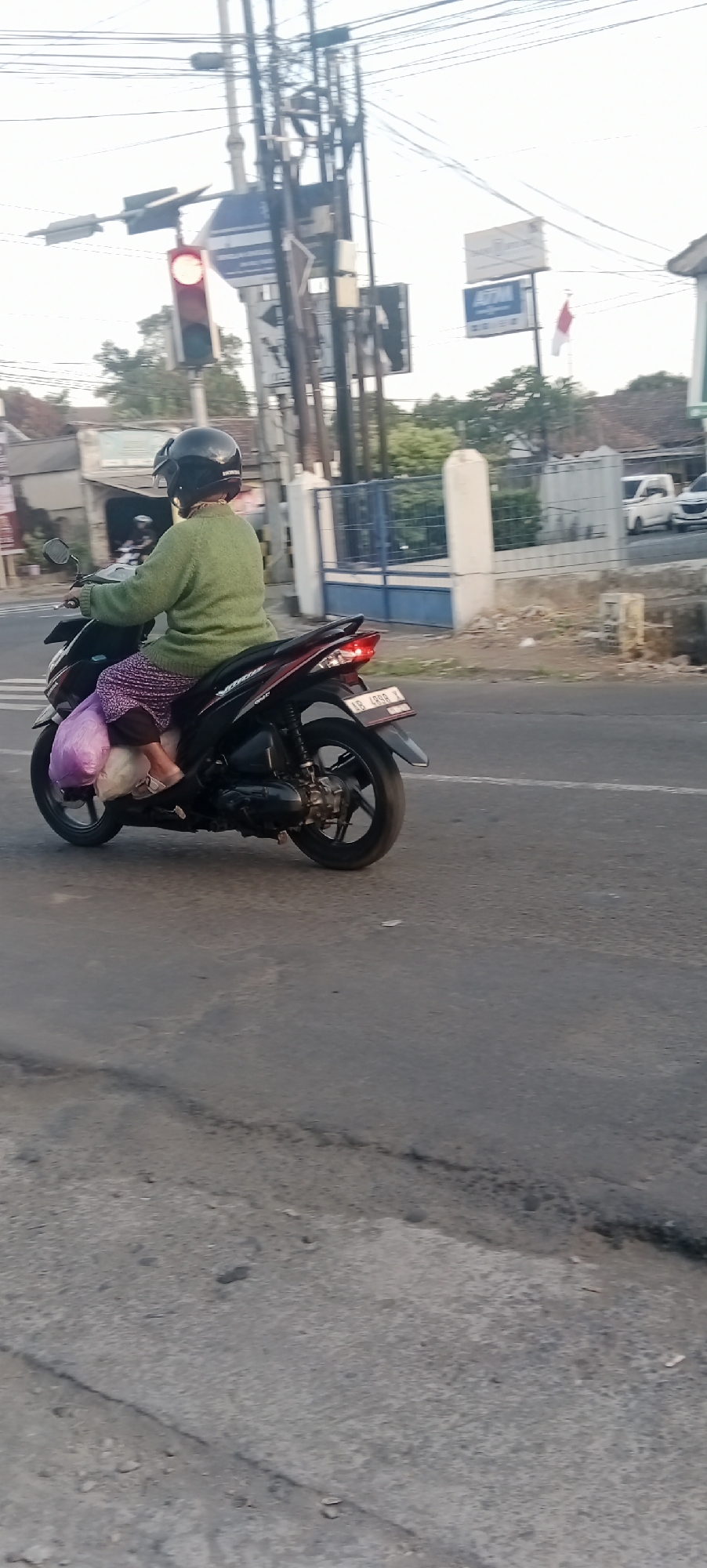 The image shows a woman riding a scooter in Maguwoharjo, Indonesia. She is wearing a green sweater and a helmet, and her scooter is carrying a purple bag. The woman is riding past a traffic light and a sign. The environment appears to be a city street with buildings and trees in the background. The road is paved and has a few cracks. The sky is cloudy.