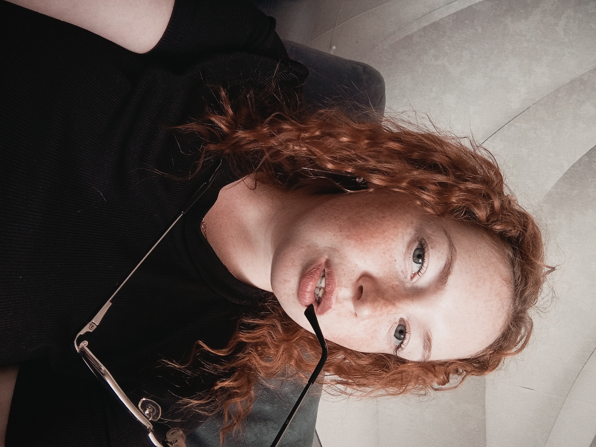 A medium-angle shot shows a woman with red, curly hair and freckled skin, appearing to be inside. She is wearing a black shirt and holding eyeglasses with a silver frame near her mouth. The background is a textured, light-colored surface, possibly a wall or ceiling. No features can be determined that would place the image in Yurgamysh, Russia.