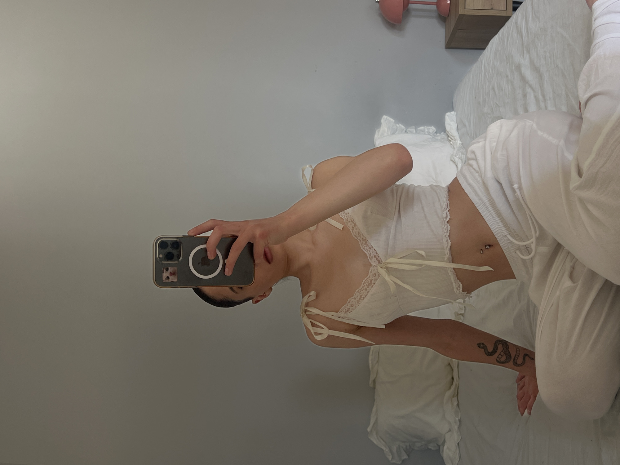 The image depicts a person sitting on a bed, holding a smartphone to take a mirror selfie. The person is wearing a white lace-trimmed camisole and white pajama pants. They have a snake tattoo on their left arm and a belly button piercing. The background shows a plain gray wall and a white bed with ruffled pillows. A pink bedside lamp is visible on the right side of the image. The photo was taken in Ternopil, Ukraine.