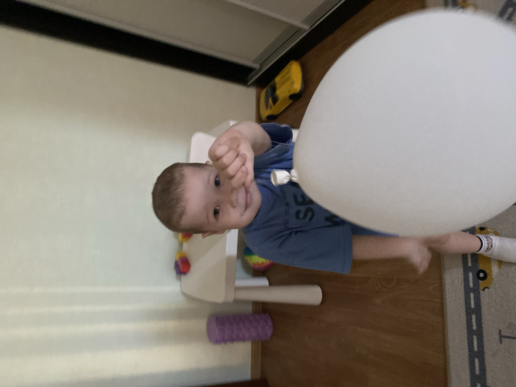 A young child is holding a large white balloon indoors. The child is wearing a blue t-shirt and has short brown hair. The child is smiling and looking at the camera, with their fist raised. The background includes a white table with a purple foam roller underneath, a yellow toy car on the floor, and part of a carpet with a car design. The environment appears to be a child's playroom or bedroom. The image was taken indoors and the location within Sochi, Russia is not discernible.