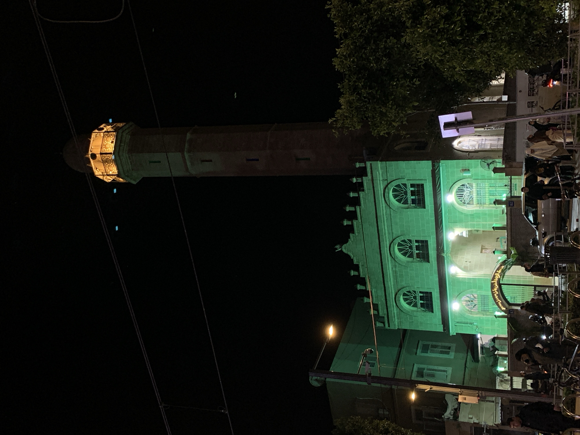 Captured at night, the image showcases a vertical composition of a building and tower in Tel Aviv, Israel.
In the foreground, multiple individuals are gathered on the street, with some appearing to be seated on bicycles.
The middle ground features a brightly illuminated green building, adorned with arched windows. Adjacent to it, a towering structure, likely a minaret, rises. The top of the minaret is illuminated with a warm light.
In the background, dense foliage and possibly more architectural elements are visible. The overall environment suggests an urban nighttime scene.