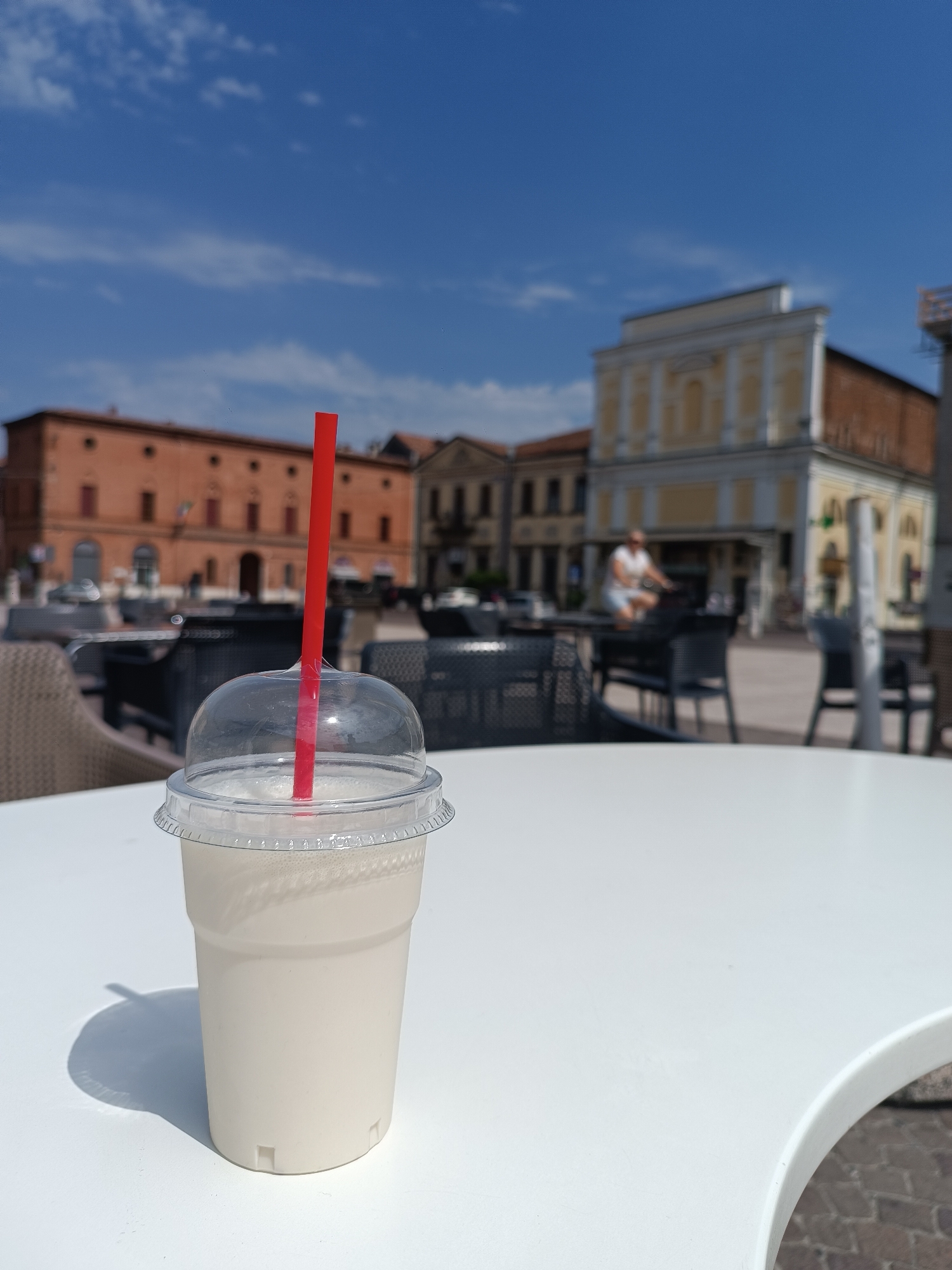 In Casalmaggiore, Italy, a light-colored beverage in a clear plastic cup with a red straw and domed lid sits on a white table. The cup casts a shadow to its left. In the background, people are visible in a plaza with several buildings. One person is riding a bicycle. The sky is blue with some clouds. The scene appears to be outdoors, possibly at a cafe.