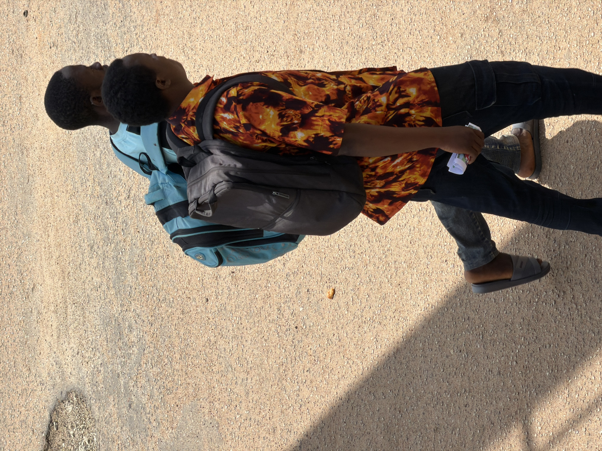 The image shows two young Black males standing side-by-side, viewed from a high angle with the camera oriented vertically. The subject on the right is closer to the camera, wearing a short-sleeved shirt with an orange, yellow, and black flame-like pattern. He has a dark backpack slung over his shoulders, and he is holding a small, folded piece of paper in his right hand. He is wearing dark blue jeans and grey slip-on sandals. The subject on the left is partially obscured by the other, wearing a light blue t-shirt and also carrying a backpack that appears to be the same style as the first. Both individuals have short, dark hair. Their faces are tilted upwards, with their eyes directed towards the sky. The setting is an outdoor, paved area, likely a street or walkway, with a light-colored, gravelly surface. The strong shadow cast across the ground indicates that the sun is high in the sky, suggesting it is midday. The overall impression is of a casual, everyday moment. There is no visible text in the image. The location context is Bukuru, Nigeria.