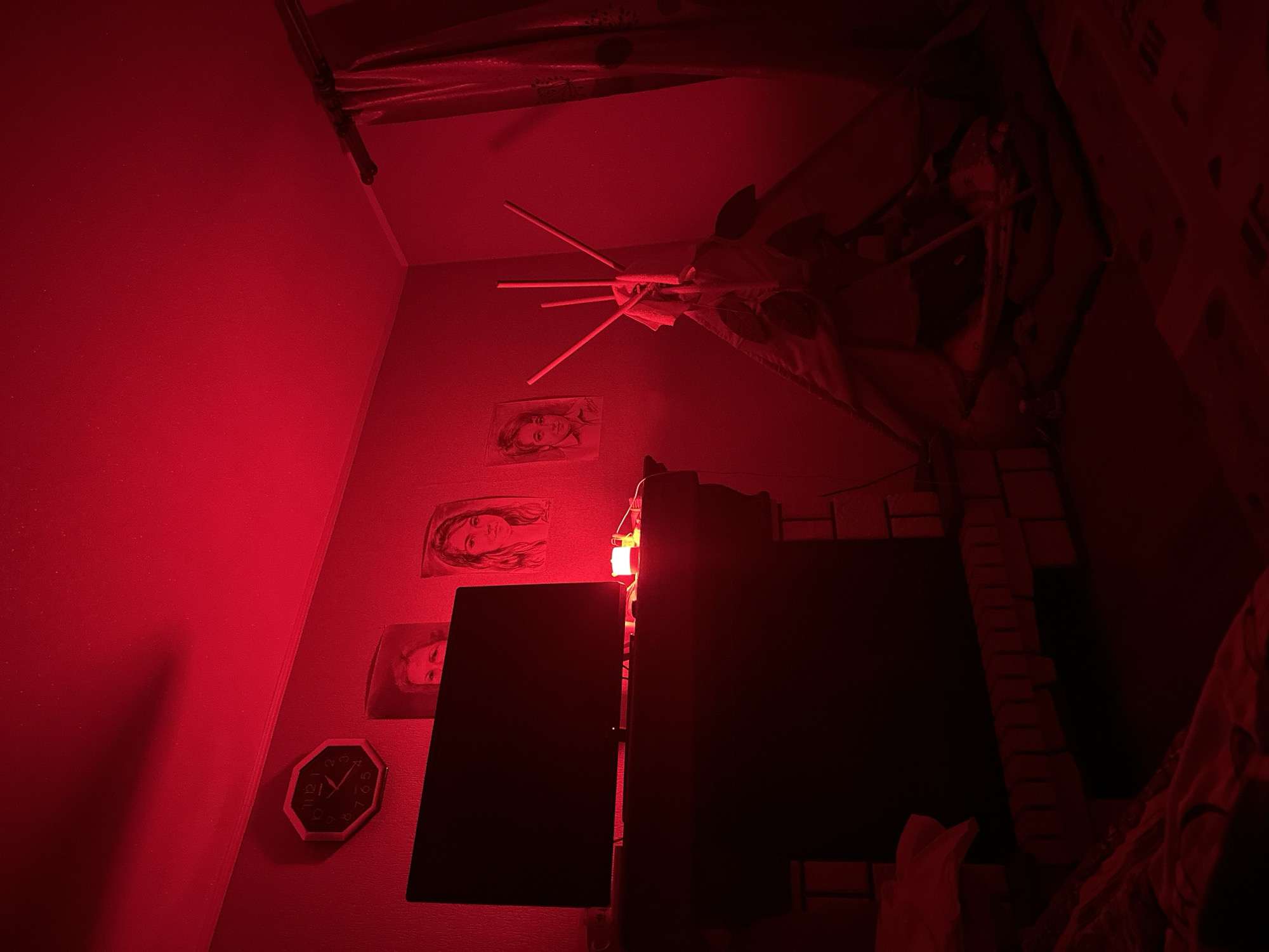 The image depicts an interior scene illuminated by red light. On the left wall, an octagonal clock is visible, indicating the time is approximately 10:25. Below the clock, a television screen is mounted above a fireplace with a brick facade. Several framed portraits of women are displayed on the wall. In the center-right of the image, a teepee-like structure is set up, with several thin wooden poles protruding from its top. A patterned curtain is visible on the far right. The scene appears to be a bedroom or living space. The image does not contain any information that would allow for localization to Nizhny Novgorod, Russia.