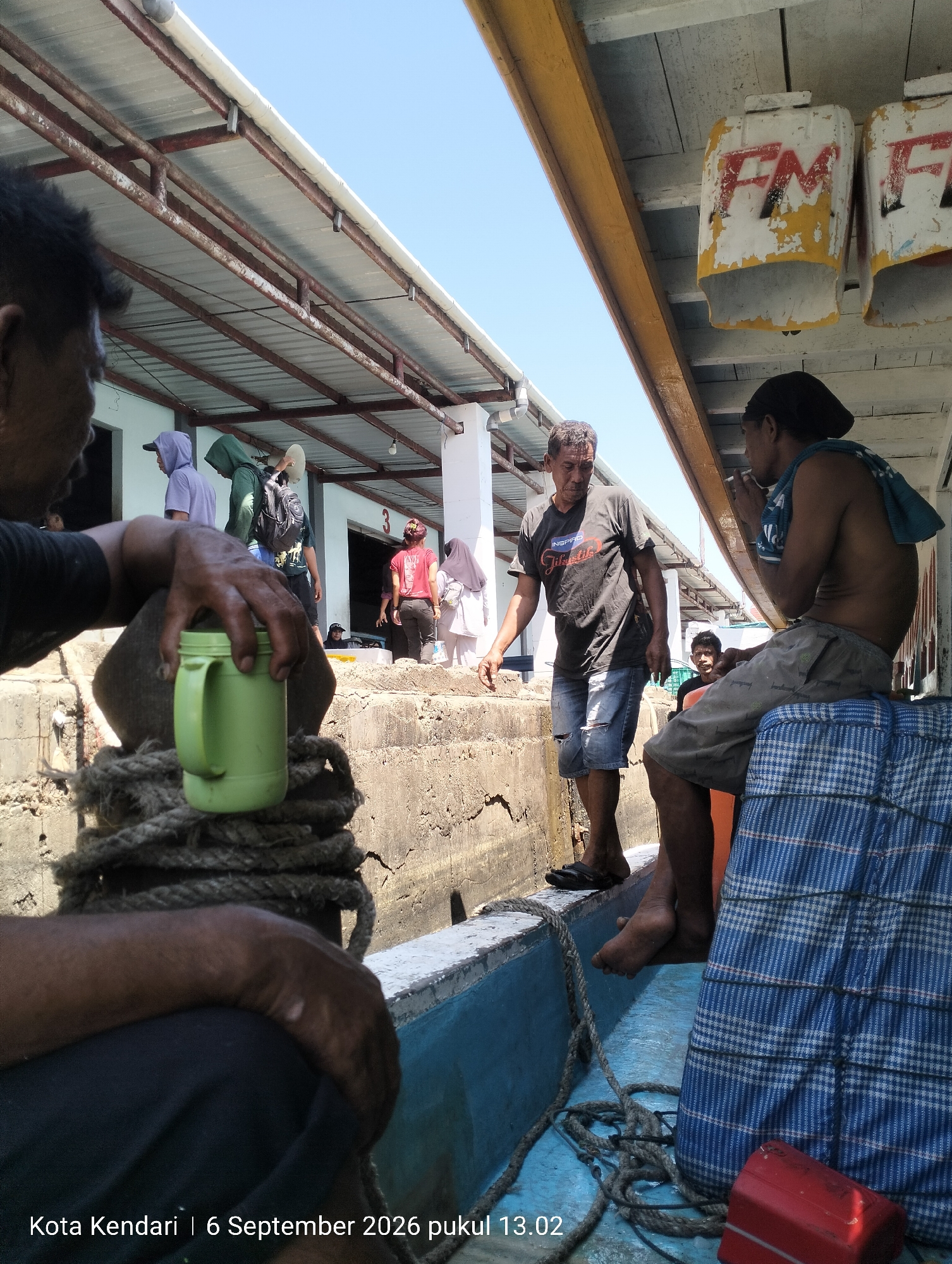 The image depicts a scene at a port in Kendari, Indonesia, on September 6, 2026, at 13:02. The foreground shows the arm and hand of a person in the lower left, resting on a coiled rope and holding a green mug. To the right, the lower body of another person is visible, seated on a blue and white checkered cloth that covers a surface, possibly a seat. The background reveals a group of people disembarking from a boat or moving along a dock. One man in a dark t-shirt and ripped denim shorts is stepping down from a higher level, appearing to assist others. Several individuals in casual clothing, including some wearing headscarves, are also present on the dock. The environment includes concrete structures, a metal roof supported by rusty beams, and clear blue sky above. There are also two weathered, cylindrical objects with faded "FM" painted on them mounted on the side of the structure. The overall impression is of a bustling port activity under a bright, sunny day.