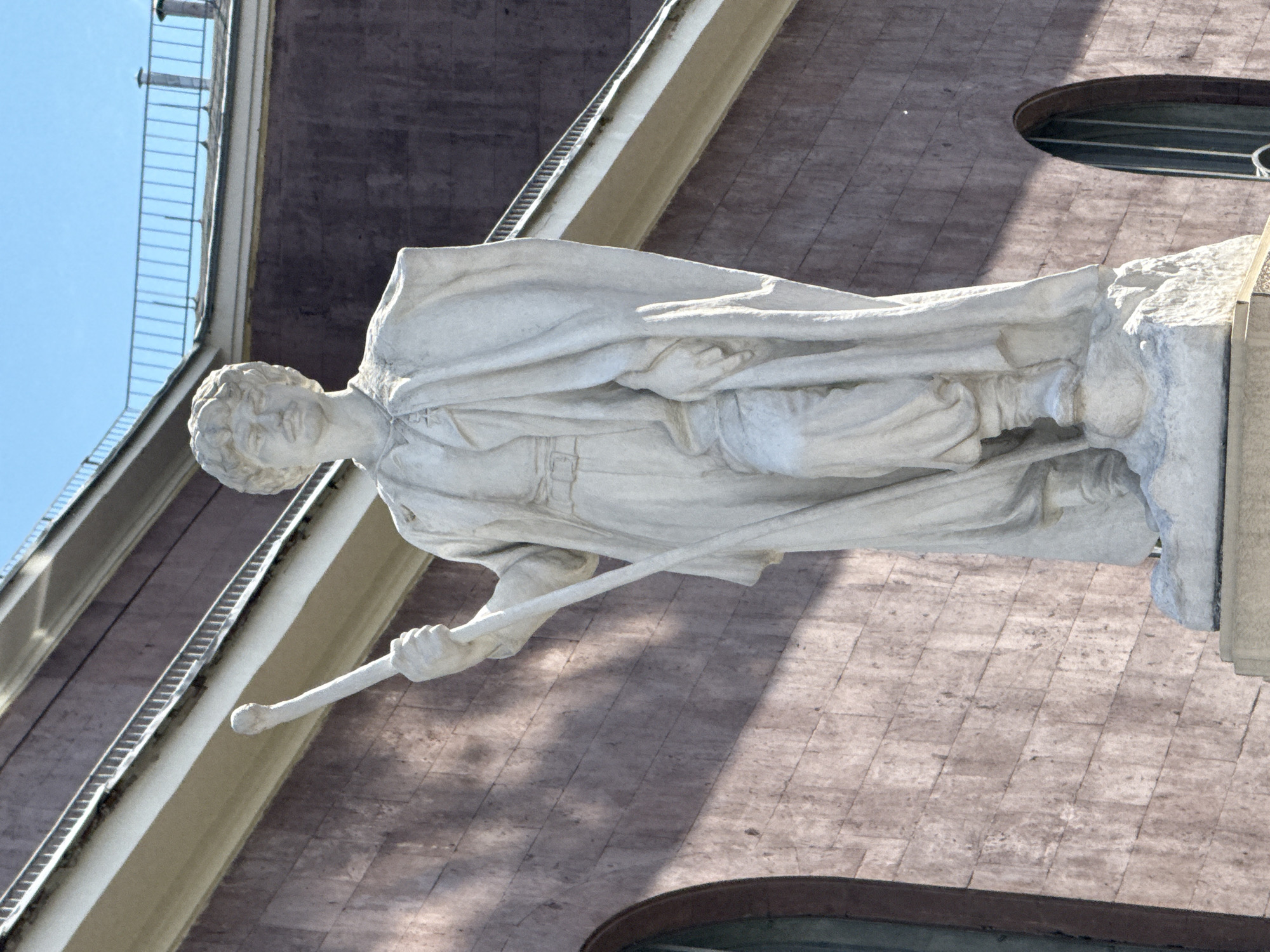 This is a vertical, close-up, eye-level shot of a white marble statue of a man, likely in an outdoor urban setting. The statue depicts a robed male figure holding a staff. The man's head is slightly tilted to his left, and his gaze is directed downwards. He appears to have short, curly hair. The drapery of his robe is detailed, with folds and textures clearly visible. His right hand grips the staff, which extends downwards and to his left.
The background consists of a building with a textured, reddish-brown facade, possibly made of brick or stone. A section of a building with a darker color and windows is visible above and to the left of the statue. A balustrade with metal railings runs along the top edge of a lighter colored architectural element that projects from the building. The sky is visible in the upper left corner, suggesting it's a clear day. The light suggests it is daytime, with the sun casting shadows on the facade of the building and on the statue itself. The overall impression is of a historic or significant architectural location. There is no visible text in the image.